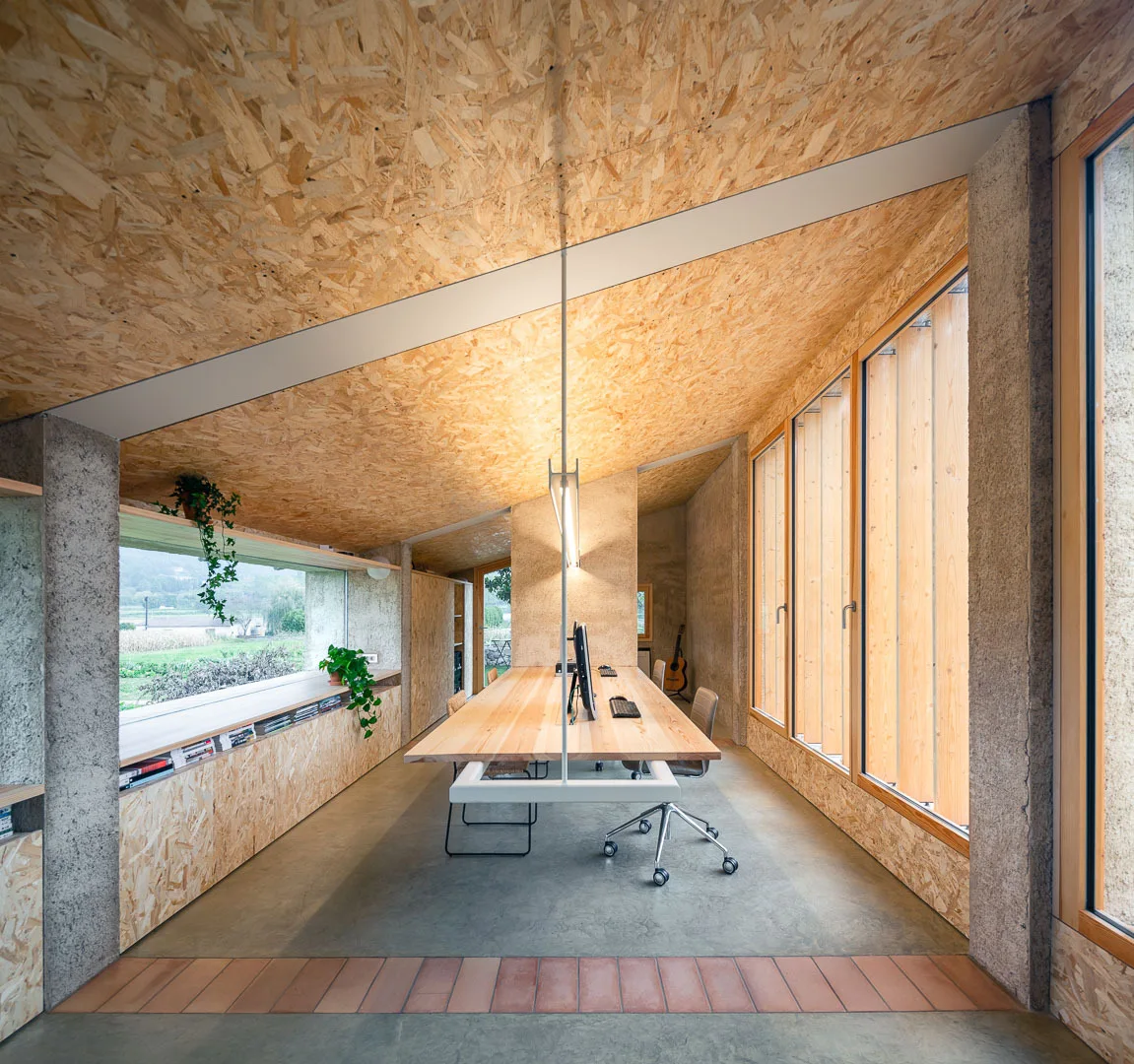 A minimalist home office features a long wooden desk, concrete floors, and walls and a slanted ceiling finished in oriented strand board.