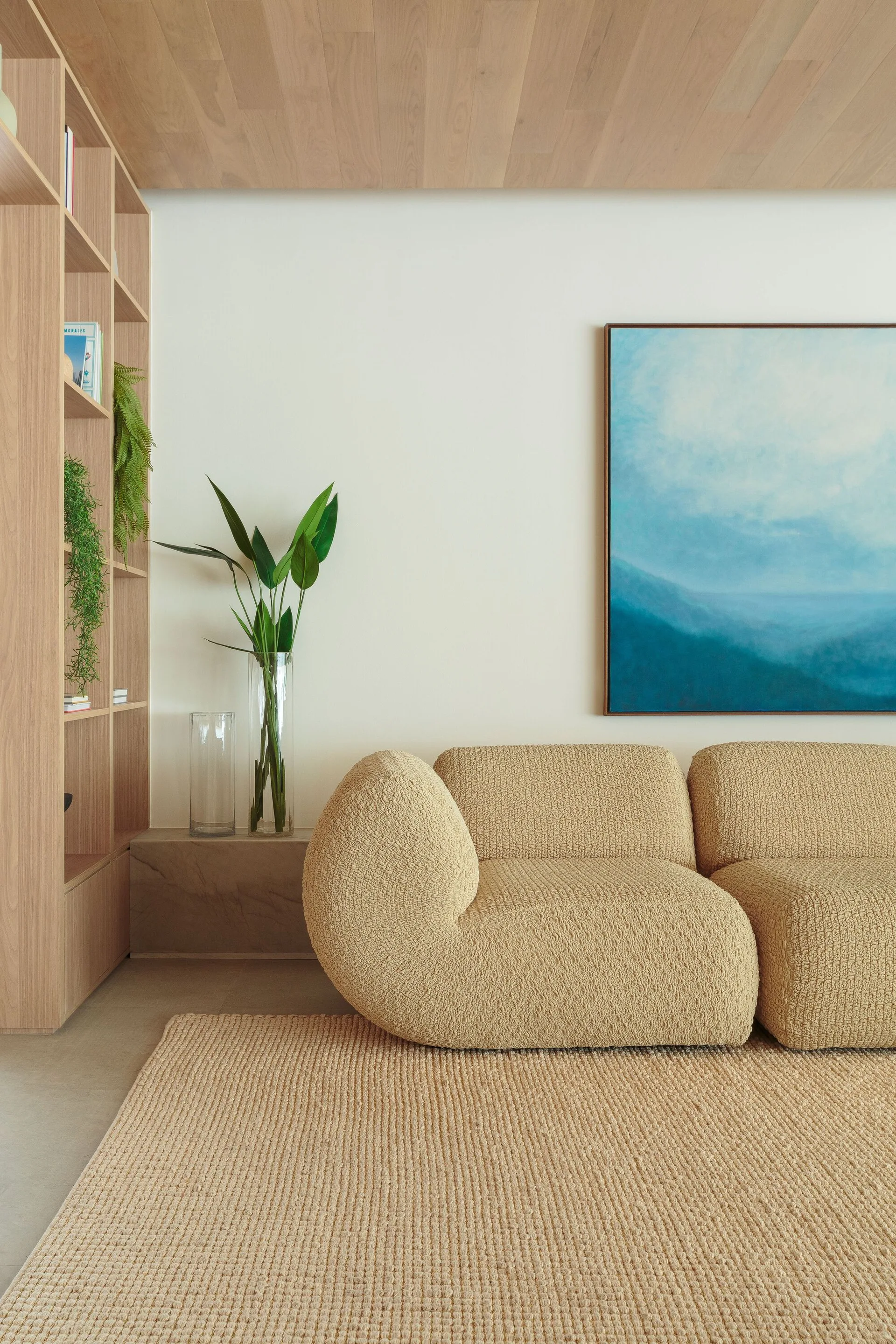 A minimalist living room features a curved beige bouclé sofa on a woven rug, a tall light wood bookcase, and a large blue painting on the wall.