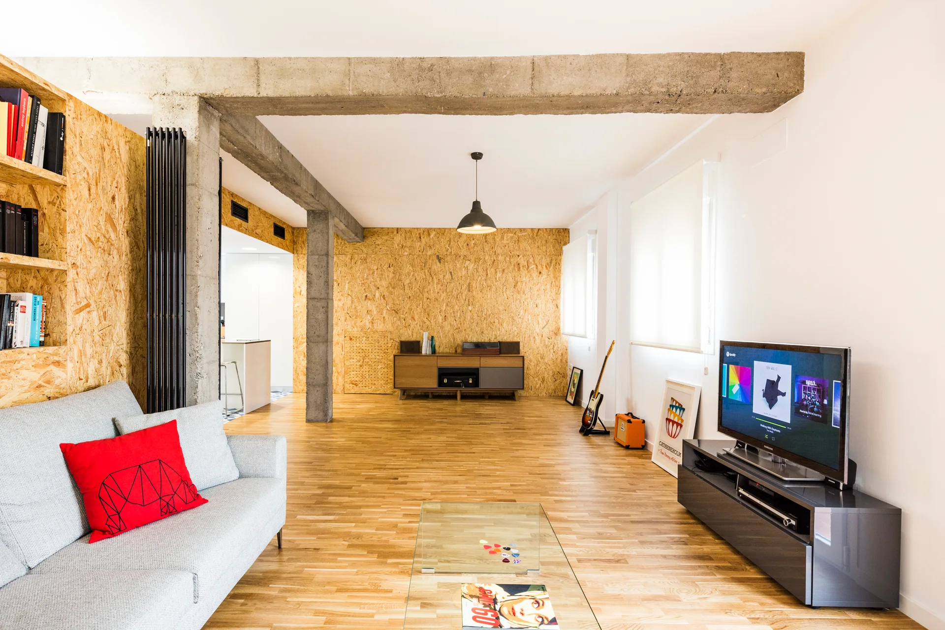 An open living room features a grey sofa, a flat screen TV, exposed concrete beams, and textured OSB wall panels.