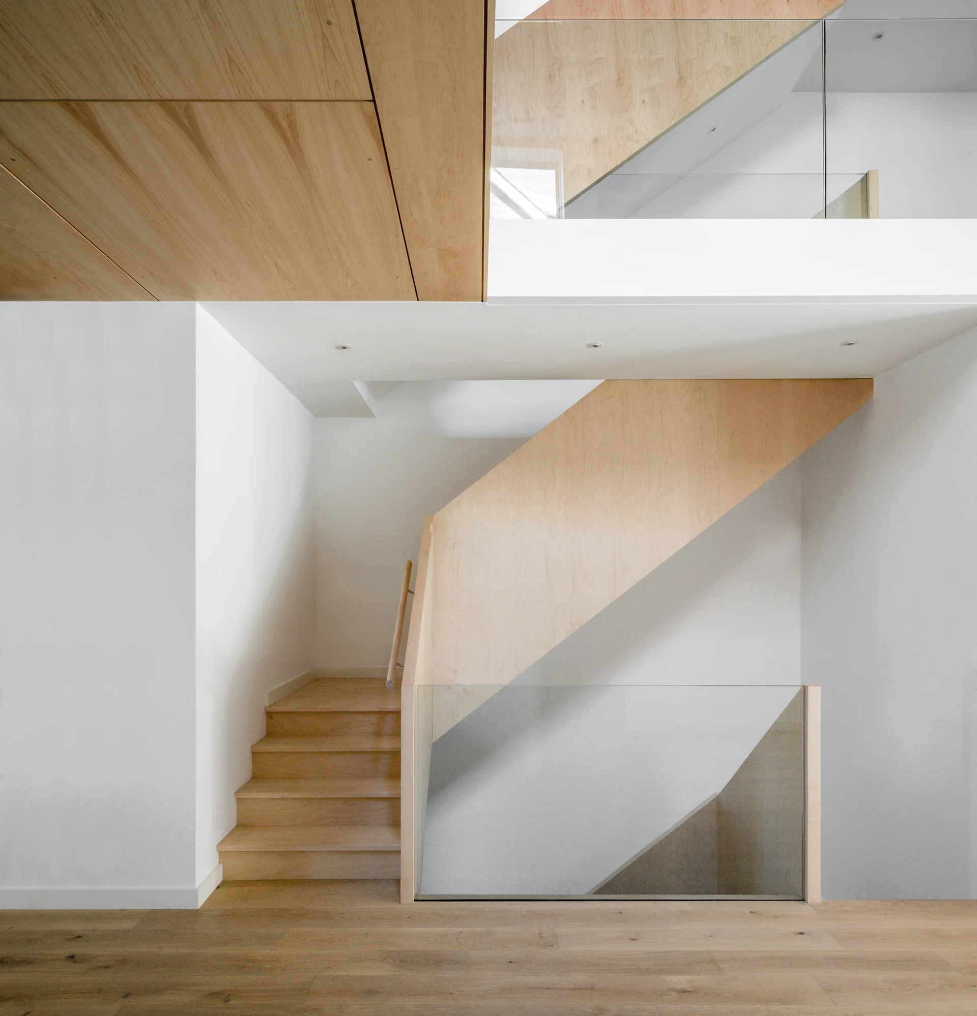A minimalist wooden staircase features clear glass safety panels, smooth white walls, and light wood grain ceiling panels.