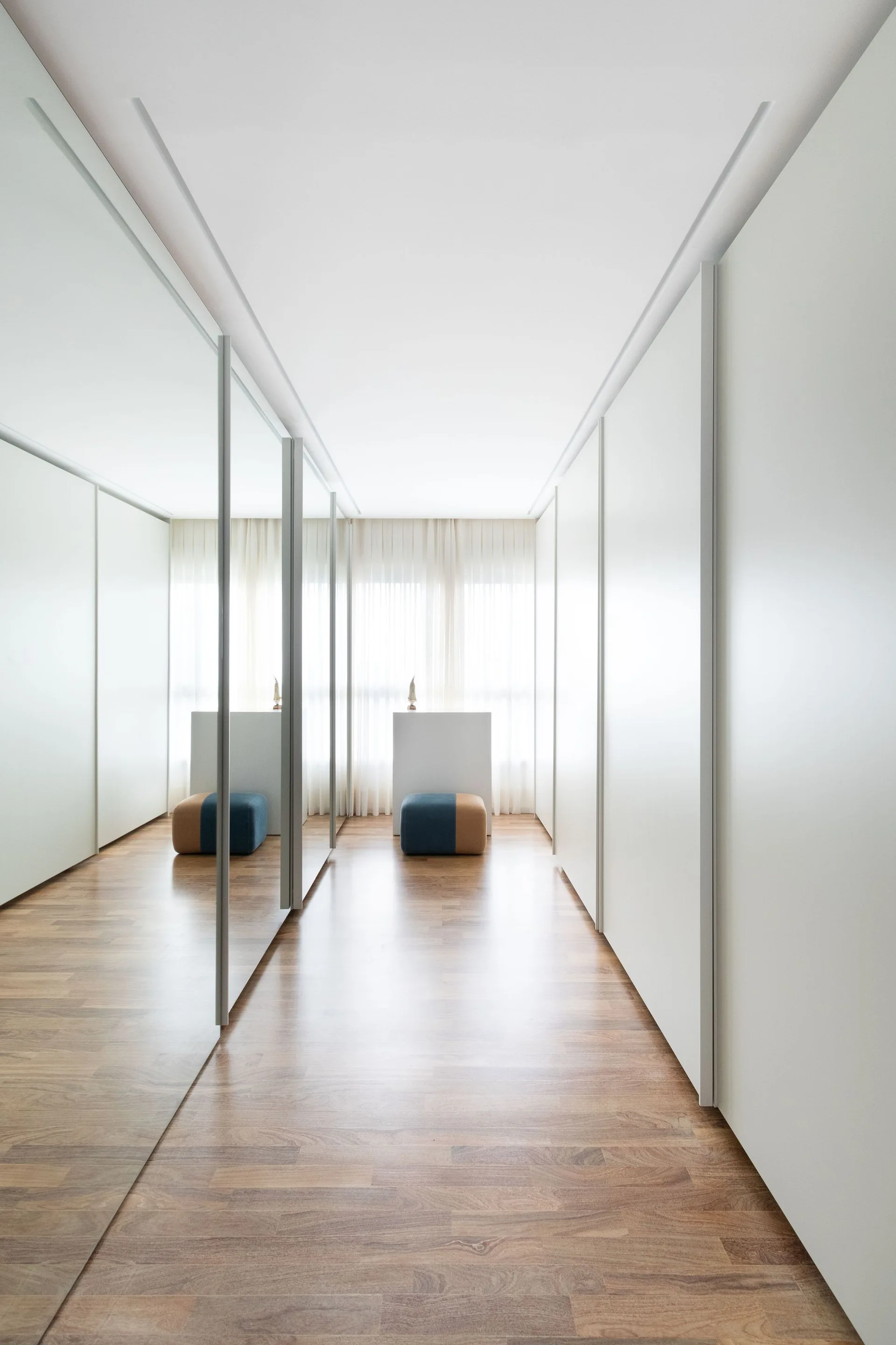 A minimalist walk-in closet corridor featuring large mirrored sliding doors, white cabinetry, warm wood floors, and sheer curtains.