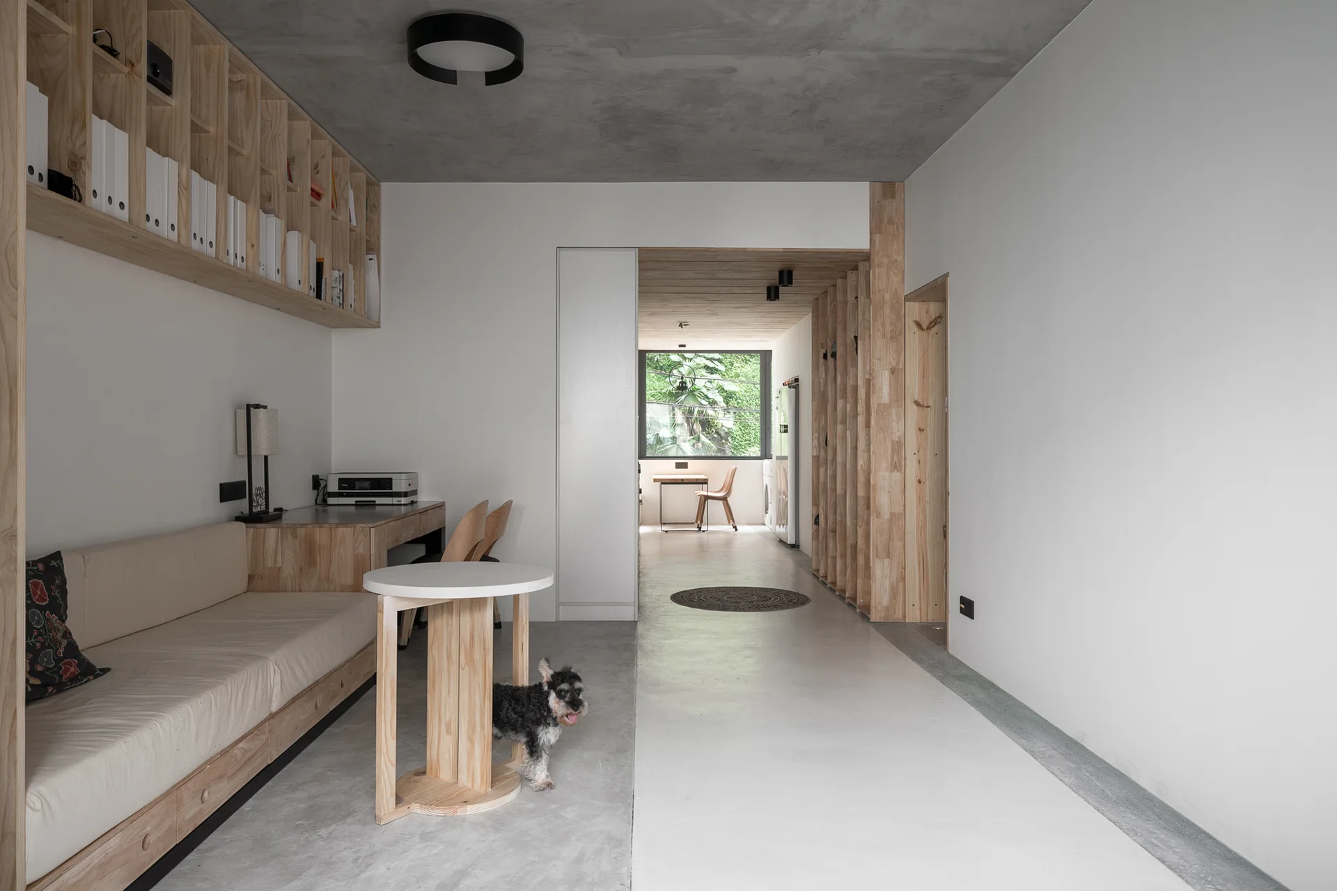 A modern room featuring a built-in wooden sofa and desk, wall-mounted wooden shelves, white walls, and an exposed concrete ceiling.