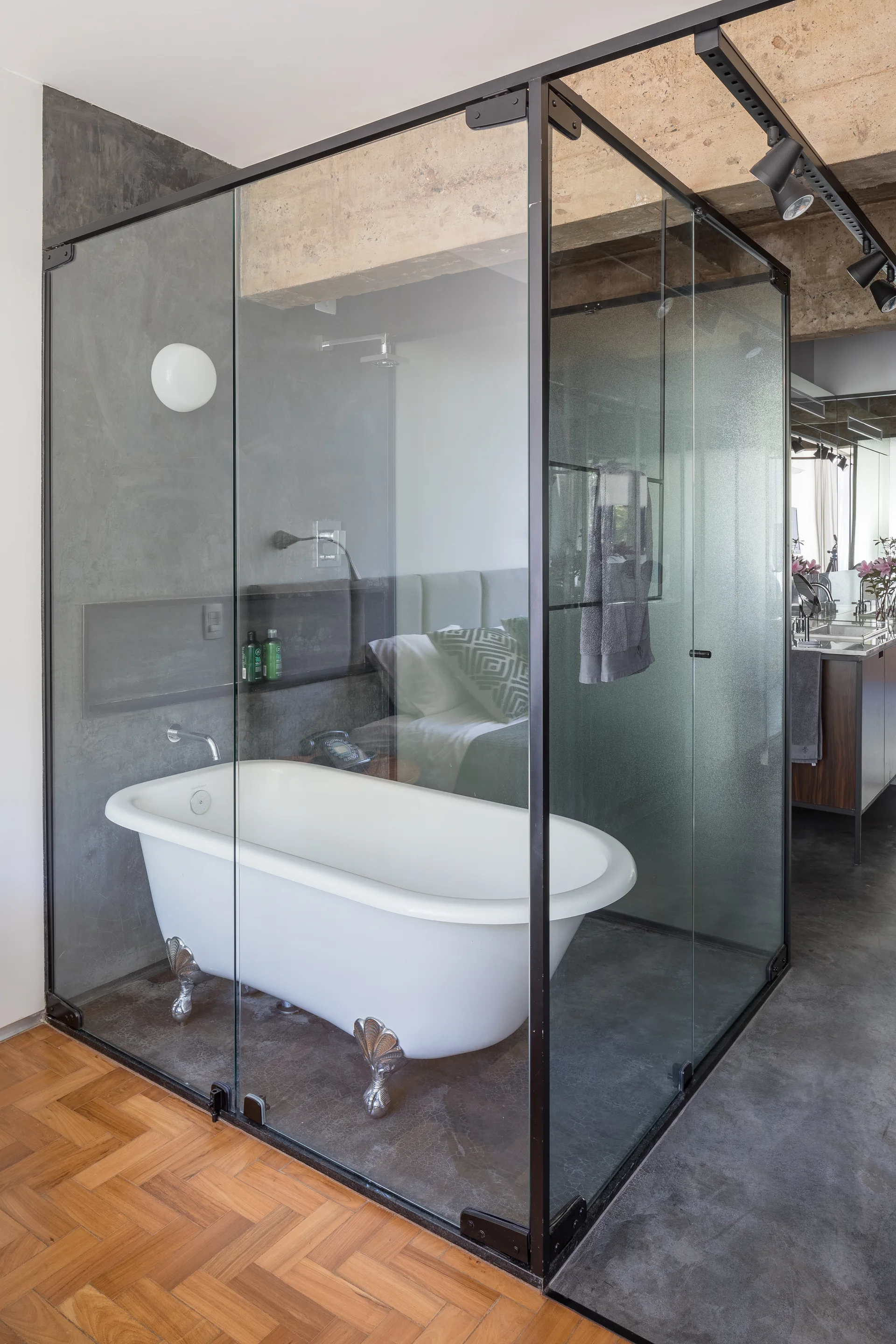 A white clawfoot bathtub sits inside a black-framed glass enclosure on a concrete floor adjacent to herringbone wood flooring.