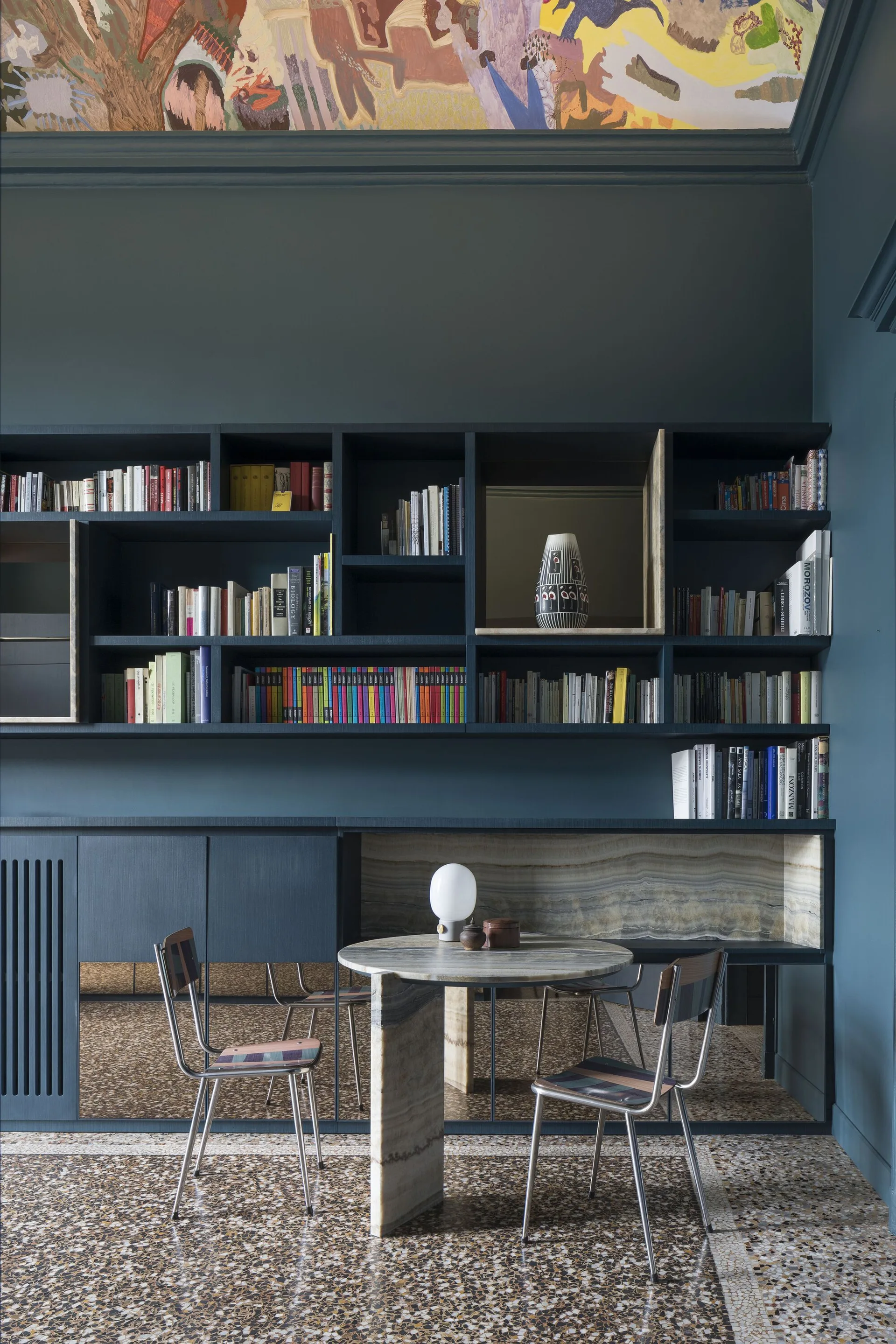 A dark blue built-in shelving unit spans a wall above a round stone table, mirrored lower cabinet fronts, and a patterned terrazzo floor.