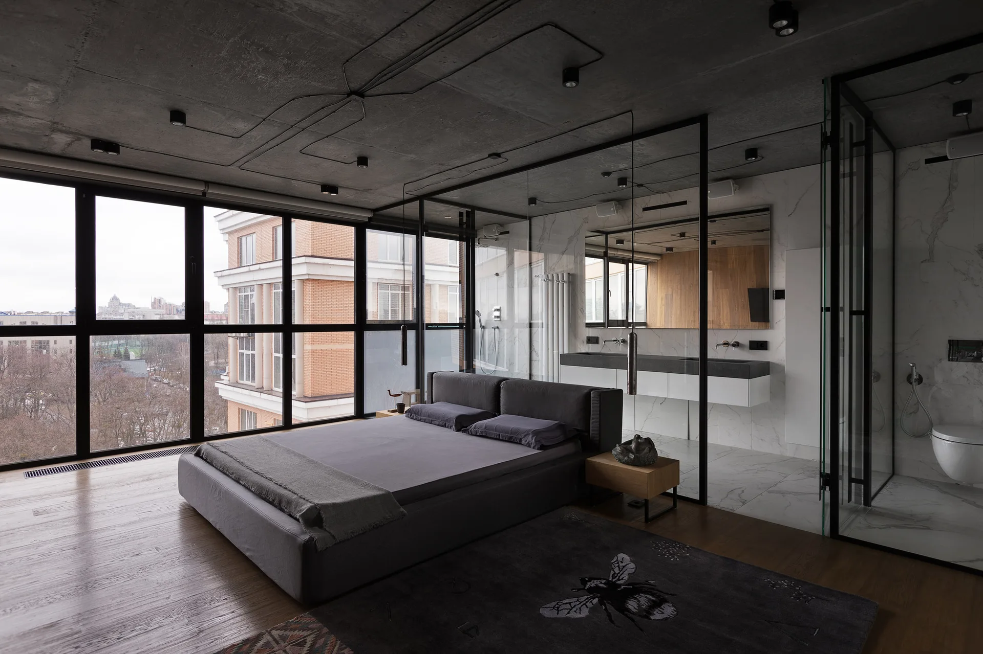 A modern bedroom with a grey platform bed, wood floors, and an adjoining bathroom separated by a black-framed glass wall partition.