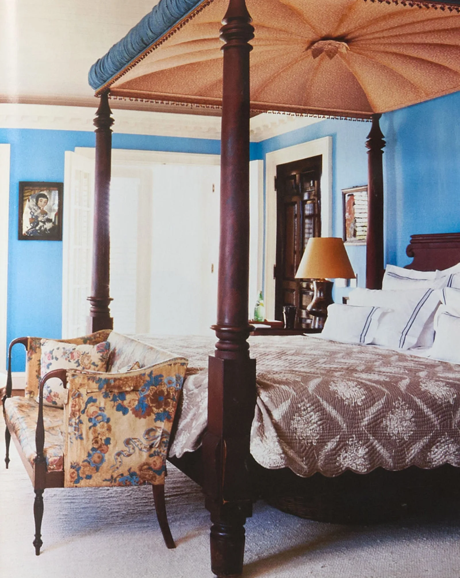 A traditional bedroom features a dark wood four-poster canopy bed set against bright blue walls with a floral-upholstered settee at the foot.