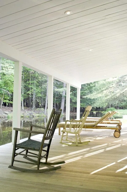 Wooden rocking chairs and sunloungers sit on a covered wood porch with a white plank ceiling overlooking a quiet pond surrounded by trees.