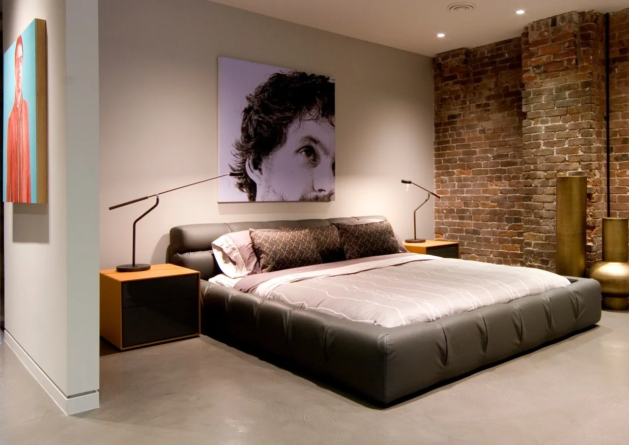 A low-profile upholstered platform bed sits between two modern nightstands with slim task lamps, set against a neutral wall and an exposed brick accent.