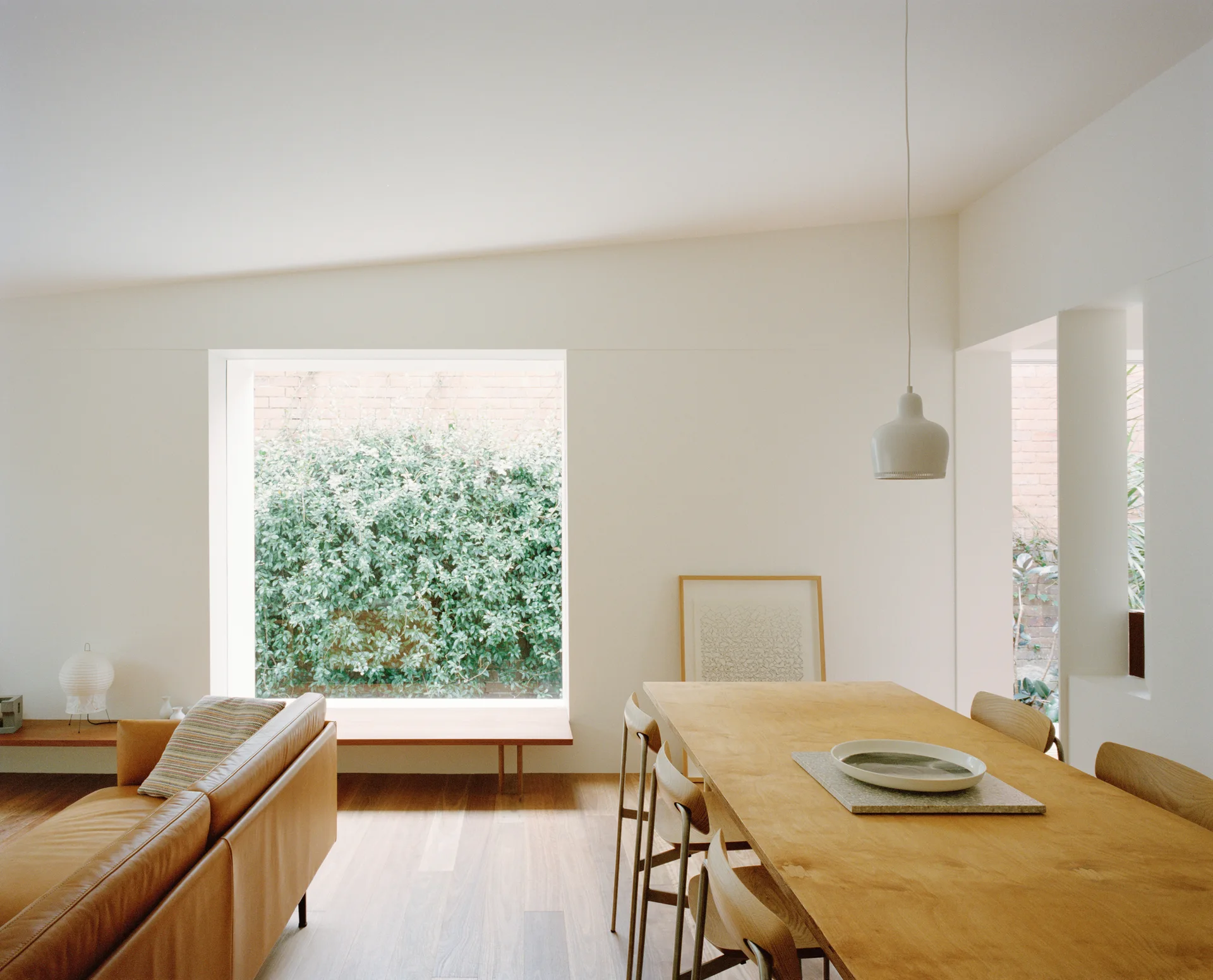A minimalist open-plan dining and living area features a wooden table and chairs, a tan leather sofa, and a large square window framing green foliage.