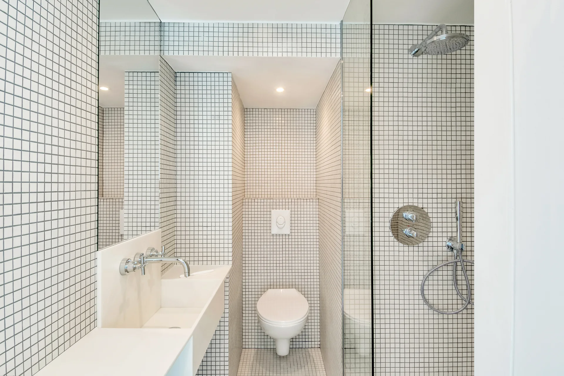 A modern white bathroom with floors and walls covered in small square grid tiles, featuring a wall-mounted sink and toilet.