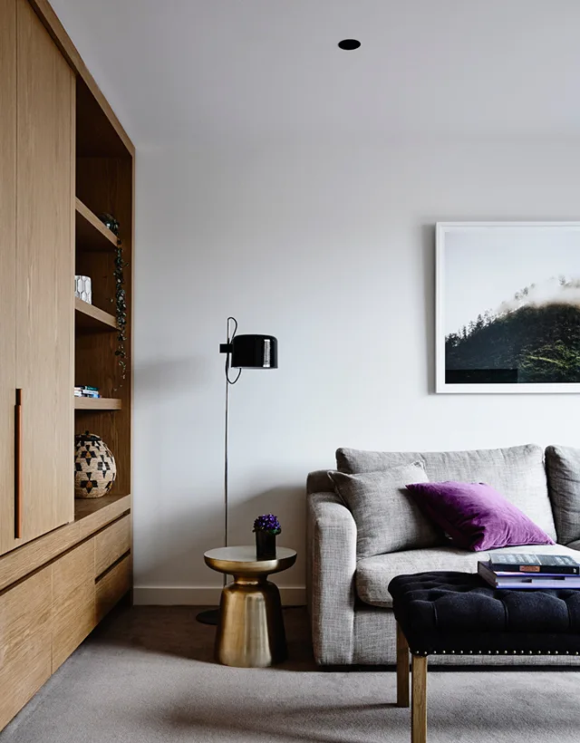A modern living room features a grey sofa with a purple pillow, integrated wood cabinetry, a gold side table, and a black floor lamp.