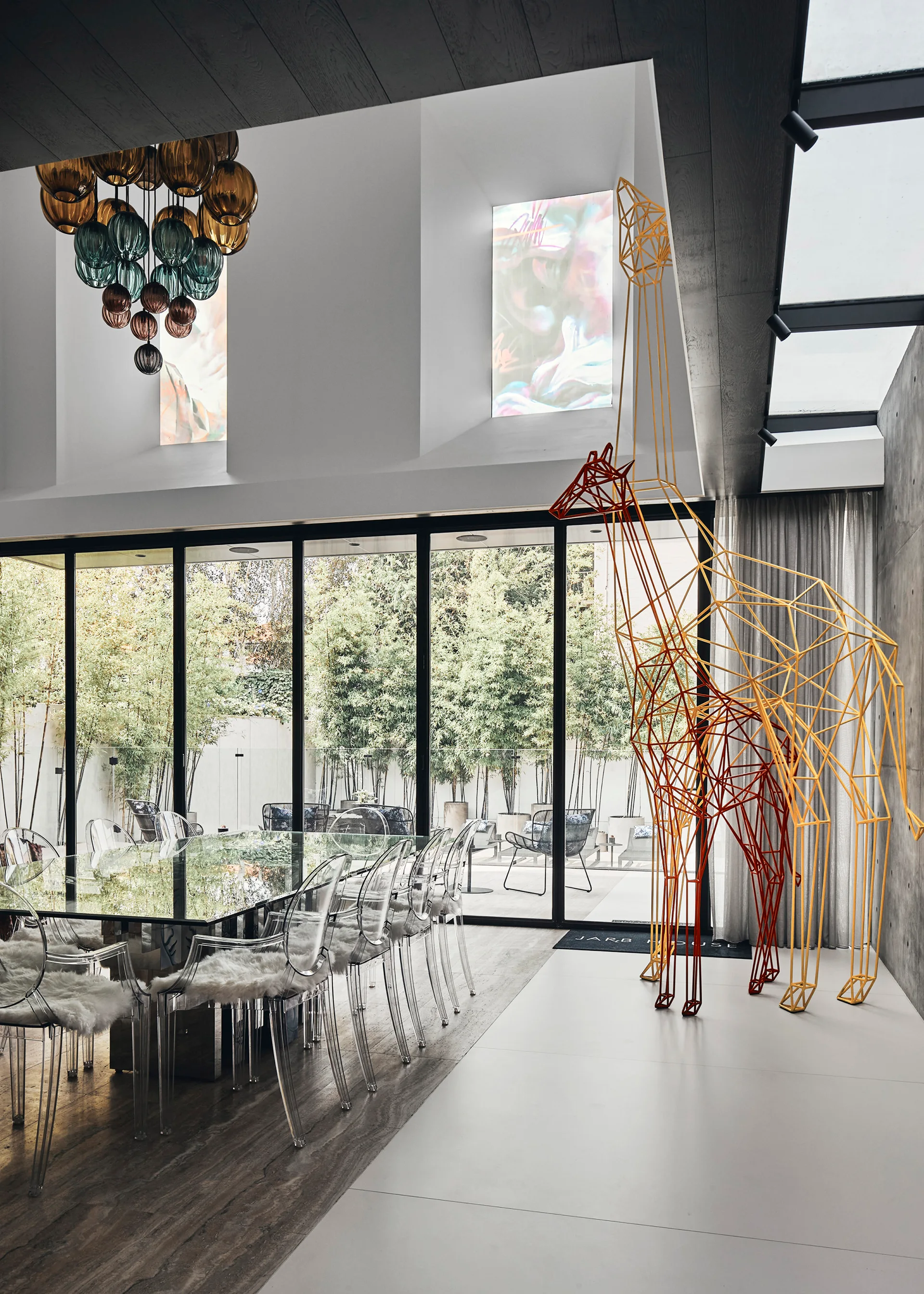 A modern dining room features a glass table with clear acrylic chairs, a multi-colored chandelier, and two tall geometric giraffe sculptures.