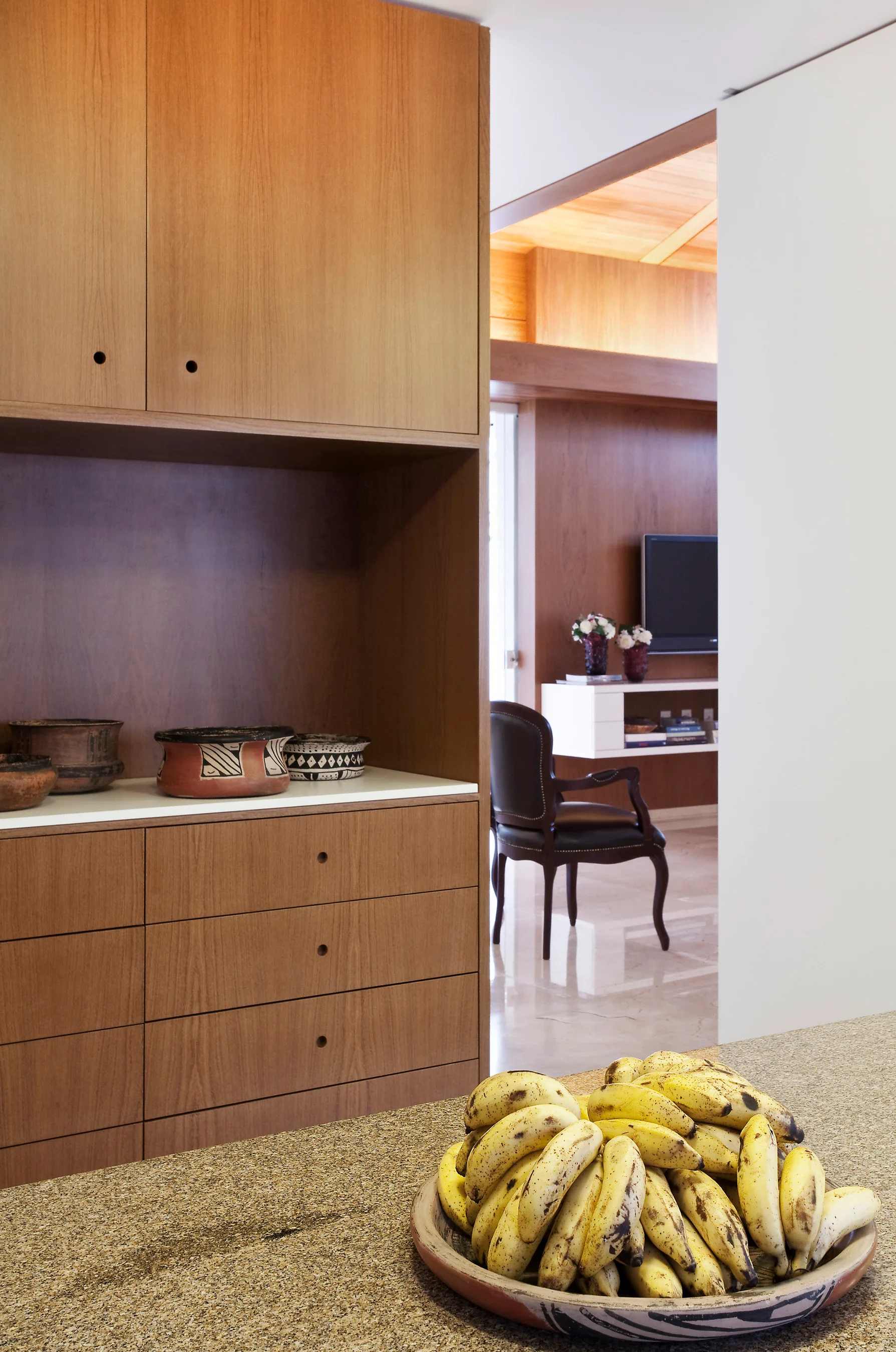 A built-in wood cabinet unit with circular pulls features an open display niche with ceramic bowls, set next to a stone-textured countertop.