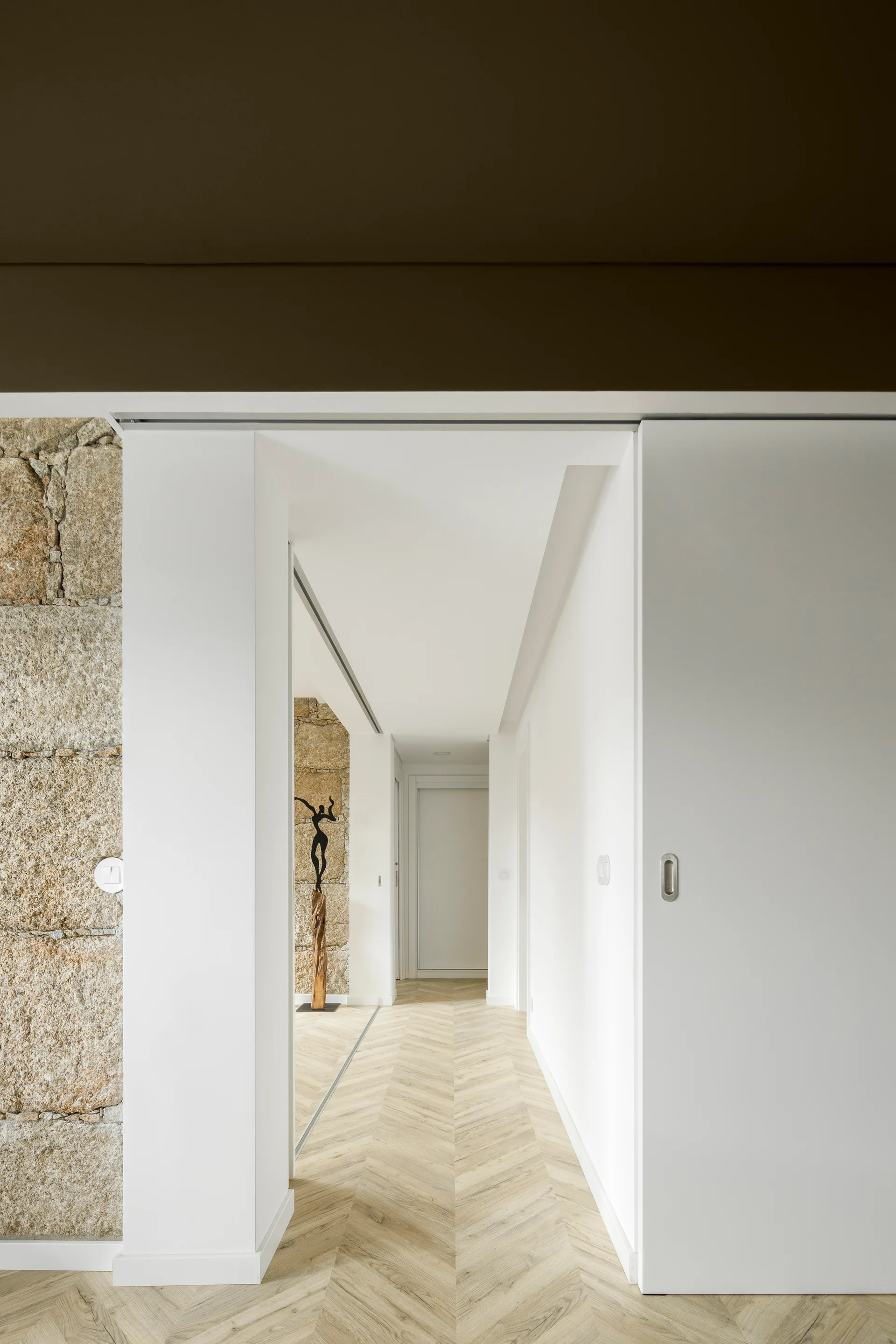 A modern white hallway features light wood herringbone flooring, a white sliding door, and an exposed rough stone accent wall.