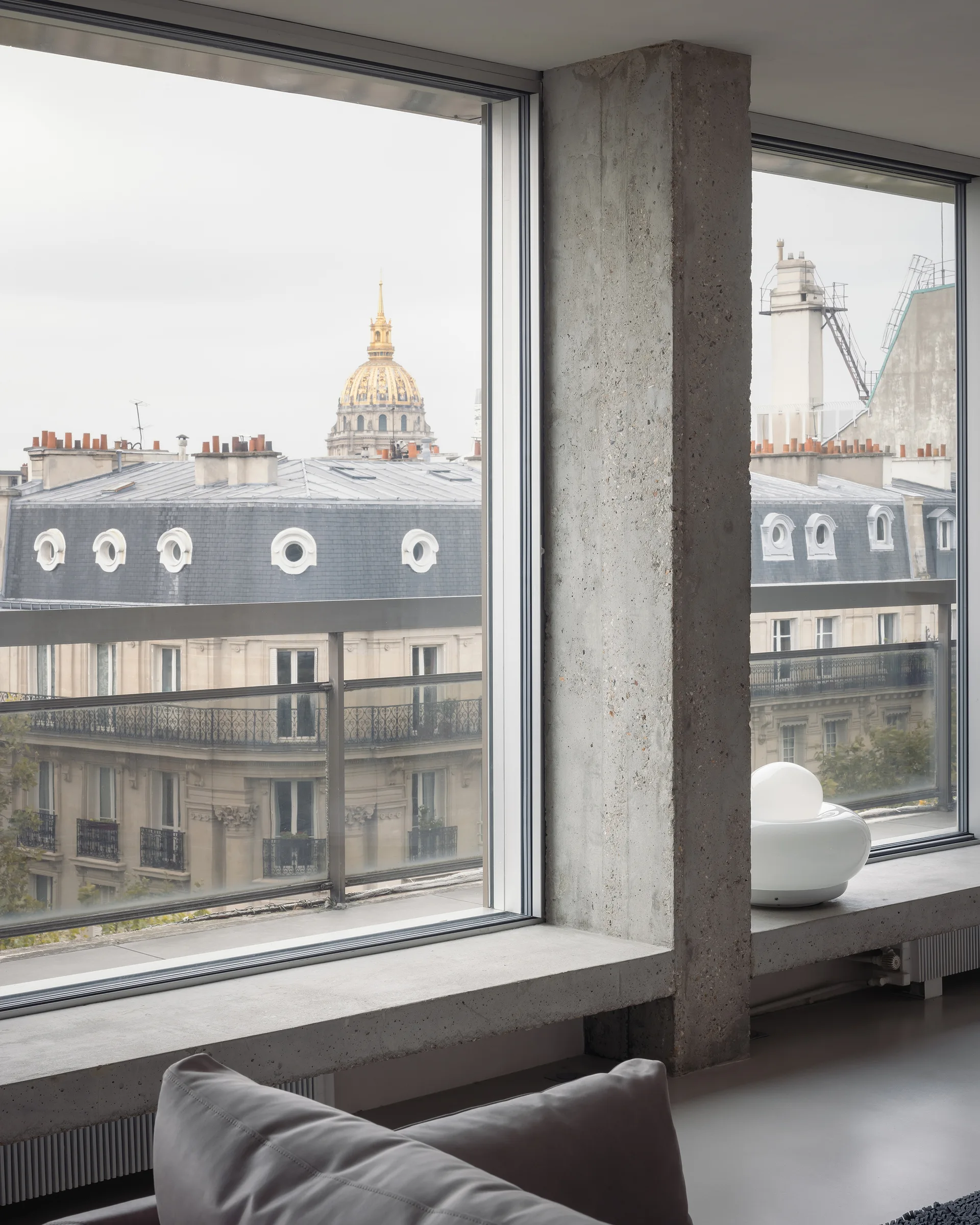 A raw concrete pillar and low ledge frame large windows overlooking classic Parisian rooftops and a golden dome.