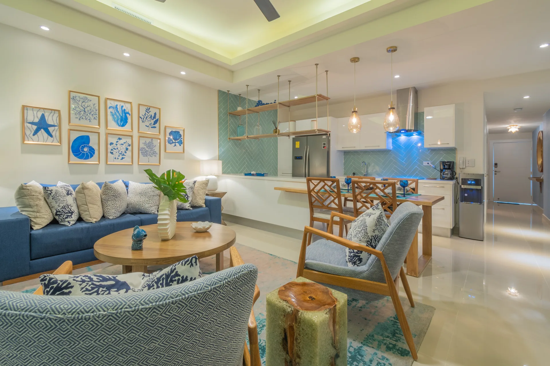 An open-concept living area and kitchen feature a blue sofa, framed botanical prints, and a teal herringbone tile backsplash behind a wooden dining table.