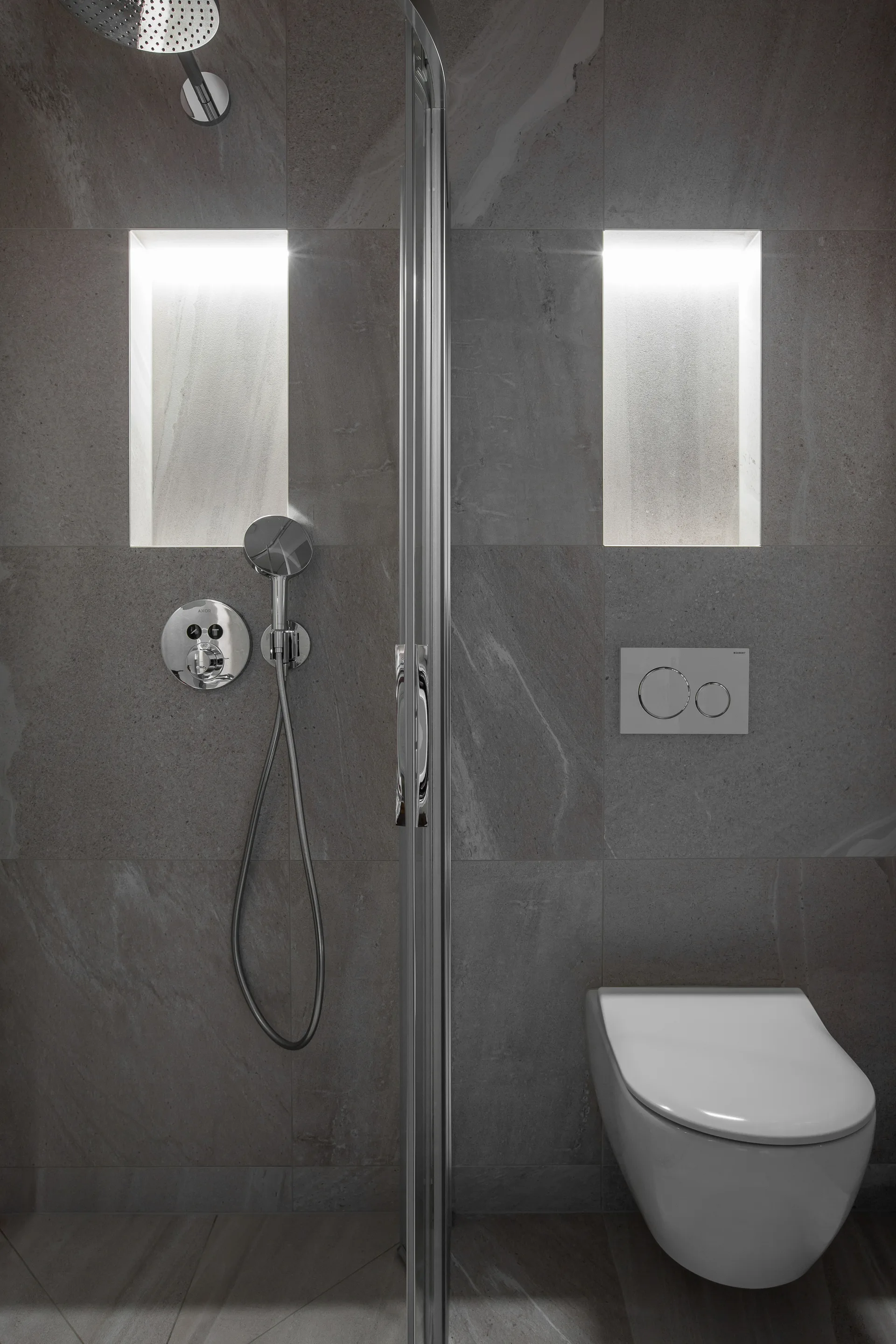 A modern grey tile bathroom featuring a glass shower partition, a wall-mounted toilet, and two vertically oriented illuminated wall niches.