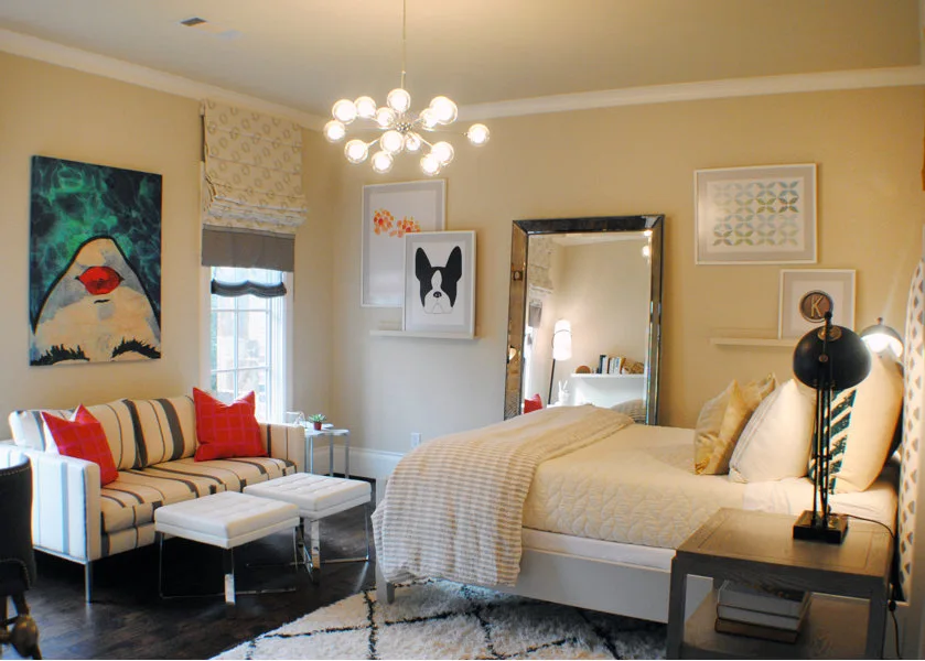 A transitional bedroom featuring a bed, a striped sofa with red throw pillows, two white ottomans, and a large floor mirror.