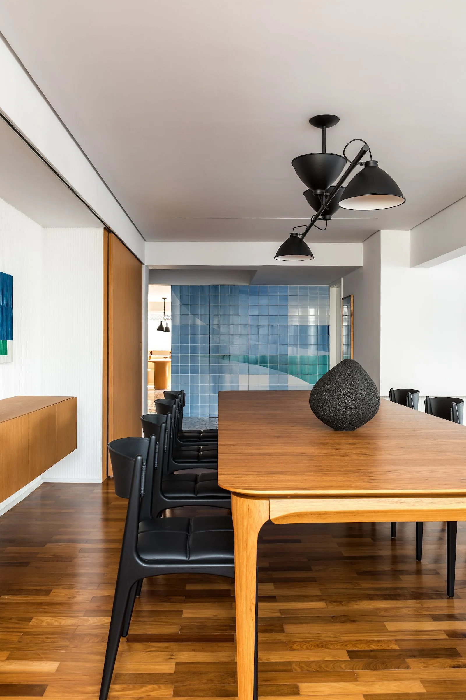 A modern dining room features a long wooden table with black leather chairs, a multi-arm black pendant light, and a blue tiled accent wall.
