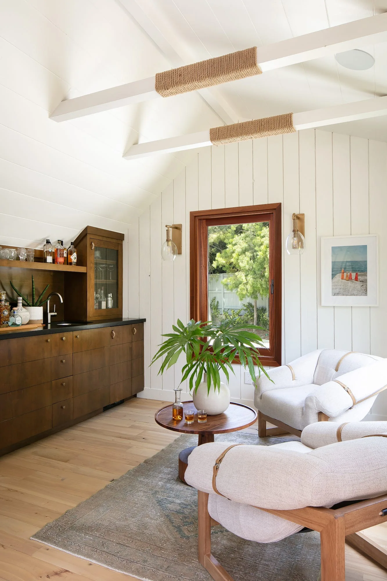 Two cream armchairs sit near a round coffee table in a lounge featuring white paneled walls, a dark wood built-in bar, and rope-wrapped ceiling beams.