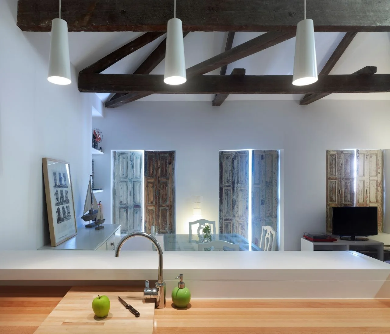 A modern white kitchen counter with a chrome faucet overlooks an open-concept room with dark exposed ceiling beams and weathered wooden shutters.