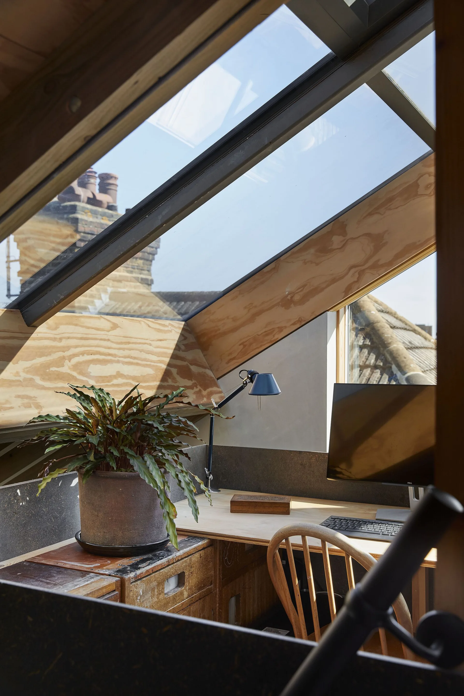 A compact home office setup under a slanted attic skylight with a wooden desk, computer monitor, and a potted plant.