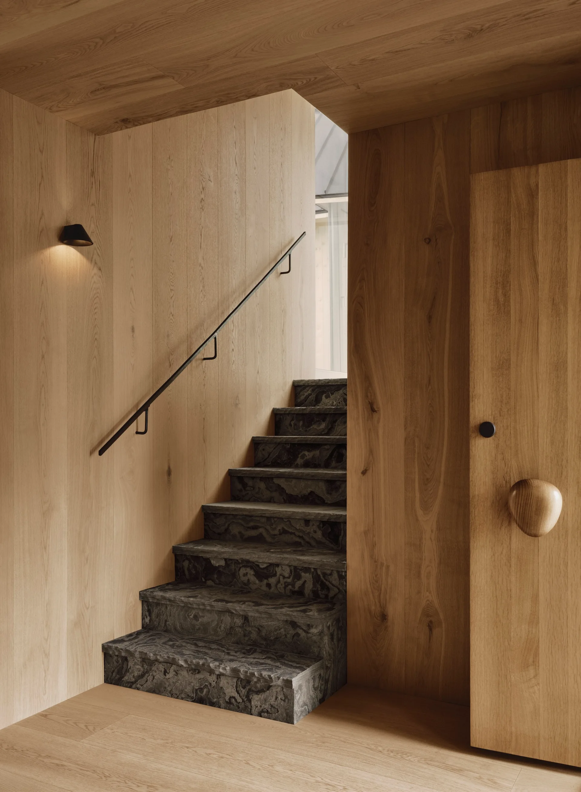 A minimalist staircase features dark veined stone steps surrounded by light wood paneled walls, a slim black metal handrail, and a small wall sconce.