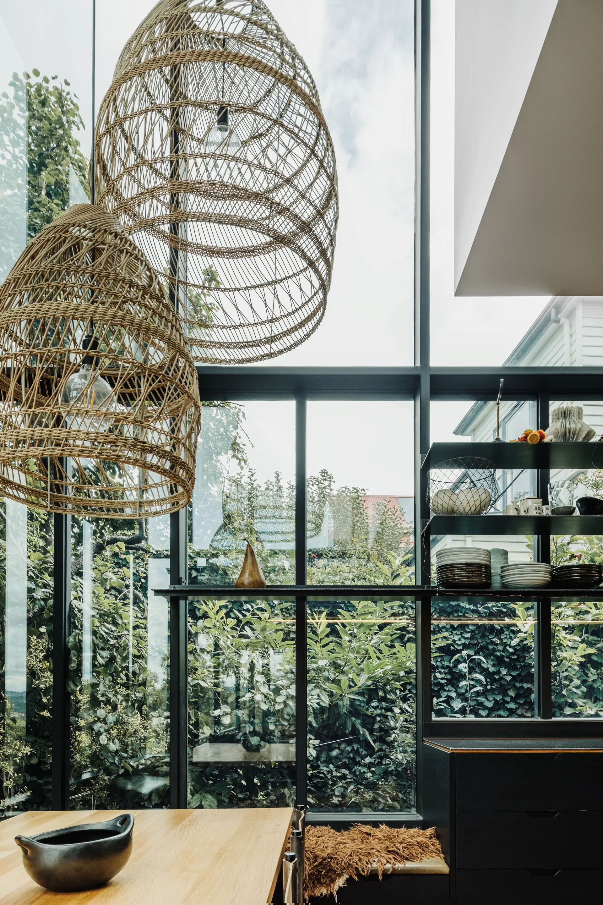 A dining space featuring two large woven rattan pendant lights hanging near a tall gridded glass window wall and black open shelving.