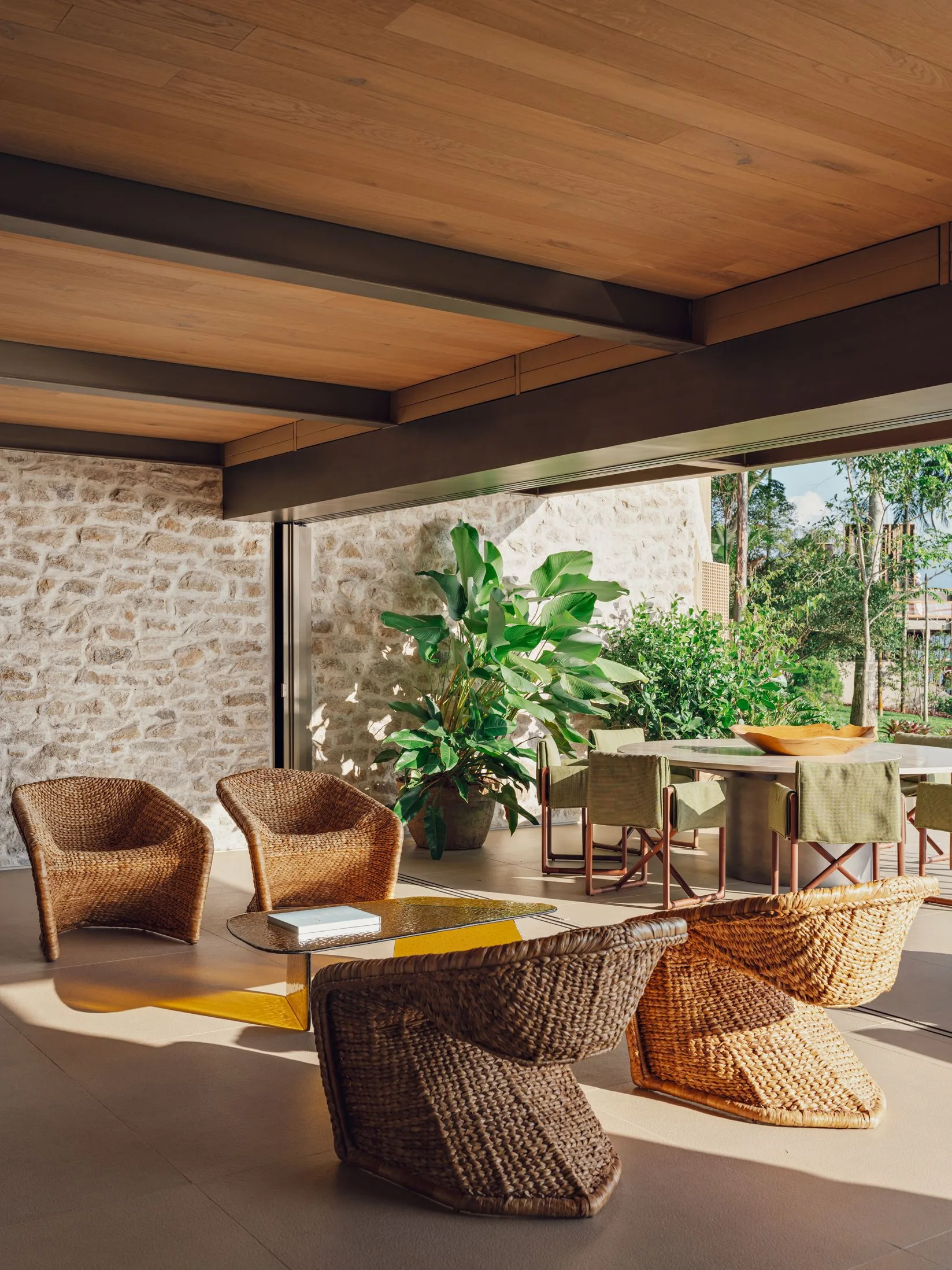A covered patio featuring woven wicker armchairs, a low coffee table, a stone accent wall, and a wood ceiling leading out to a garden.
