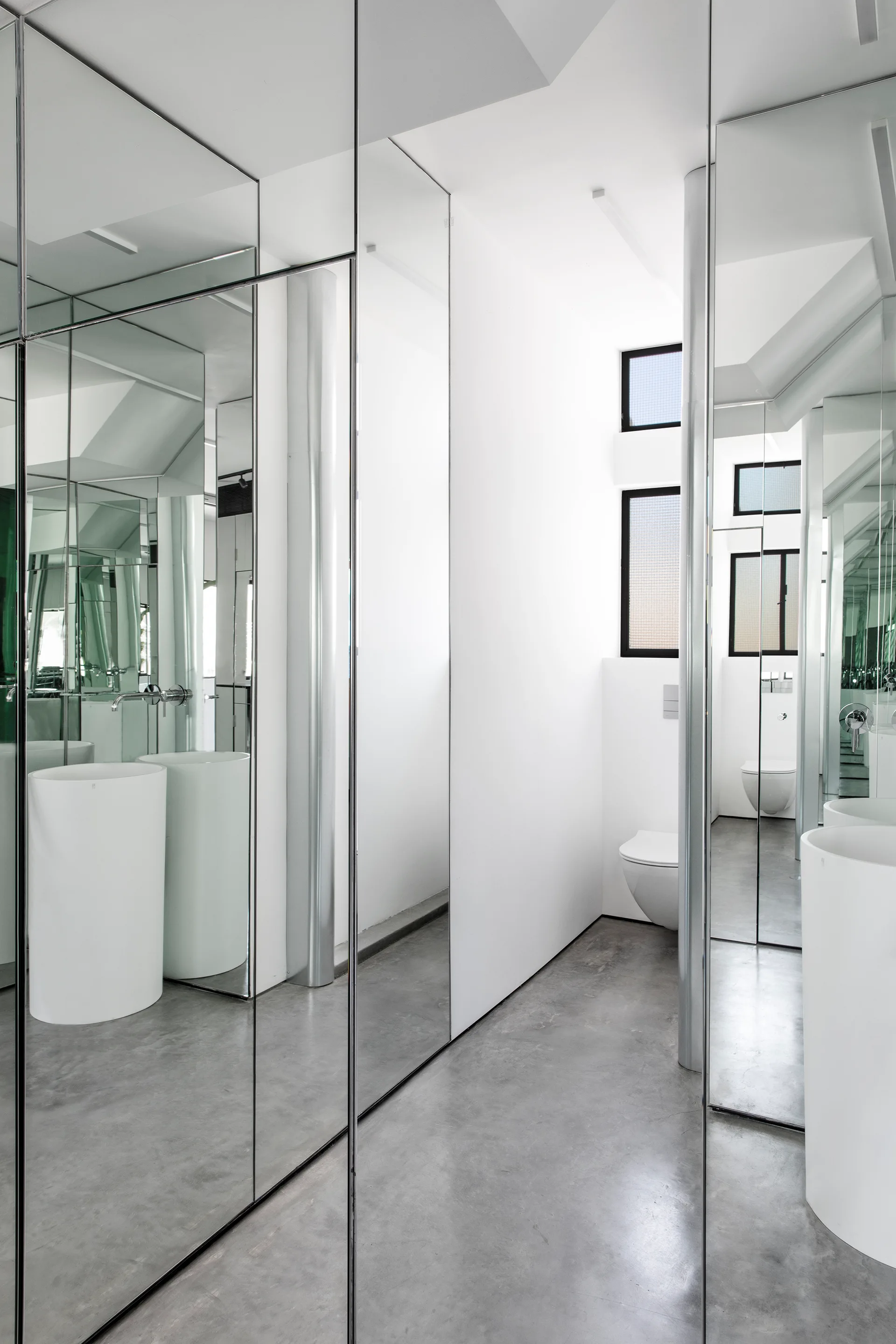 A minimalist bathroom featuring floor-to-ceiling mirror panels, a white cylindrical pedestal sink, a wall-mounted toilet, and grey concrete floors.