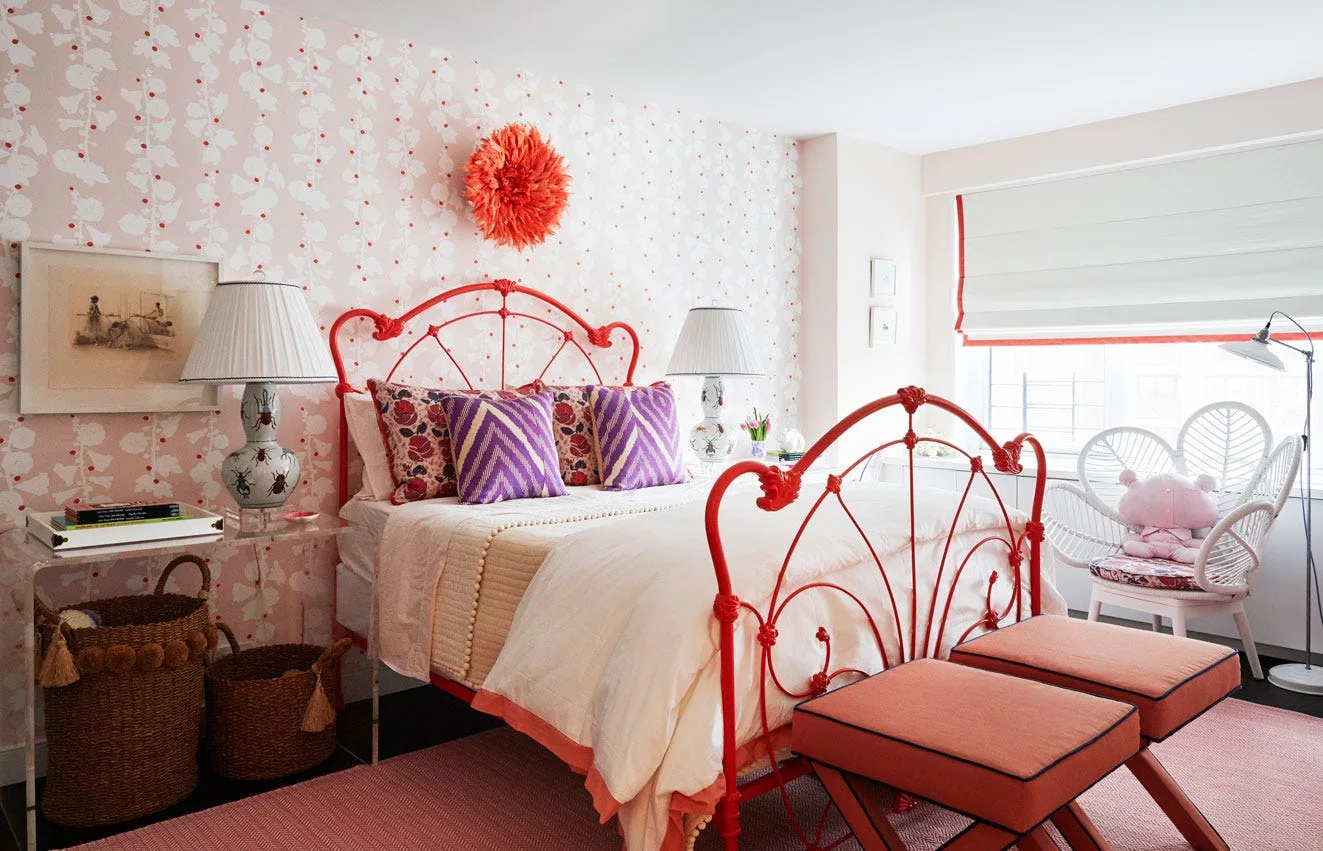 An eclectic bedroom features a bright orange metal bed frame against pink floral wallpaper, flanked by table lamps and an acrylic nightstand.