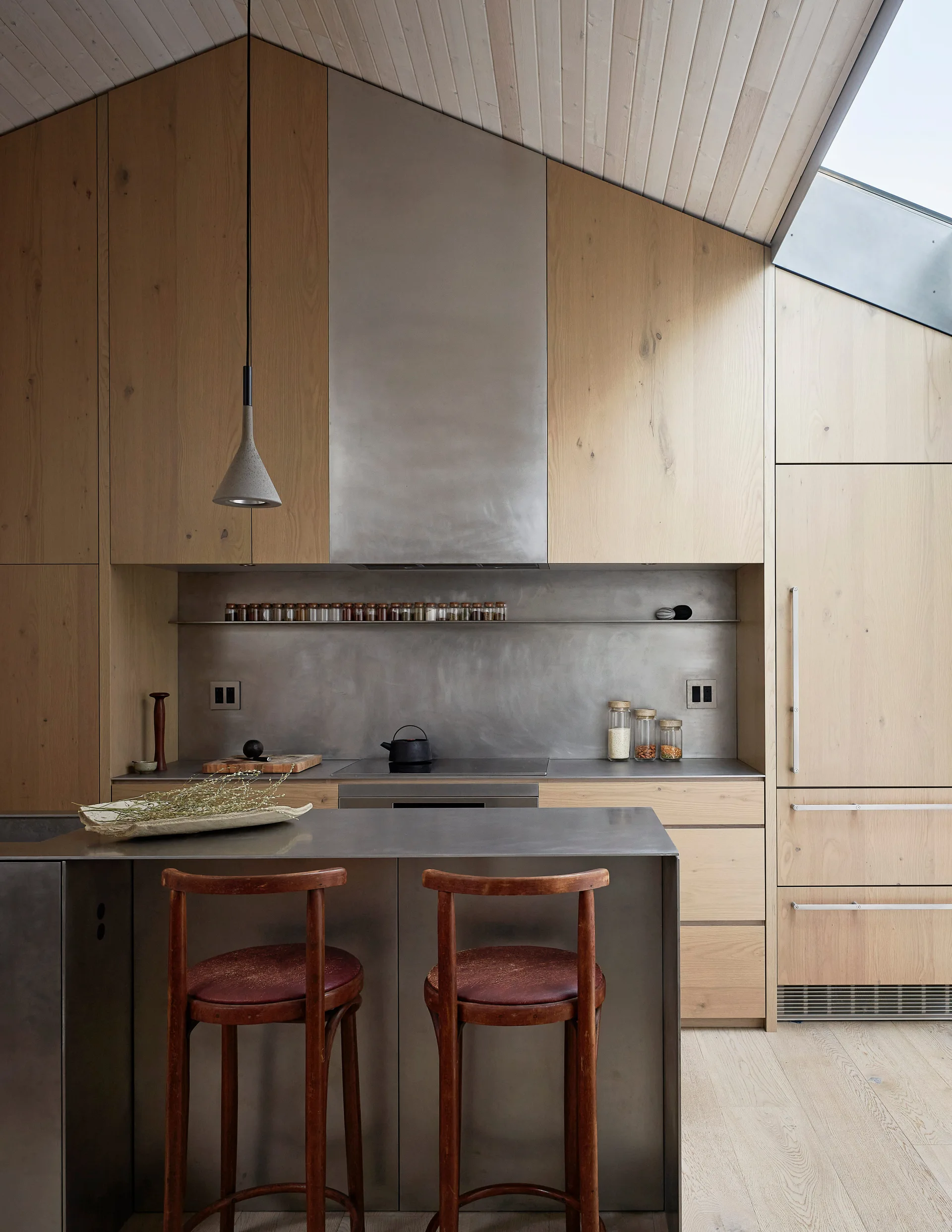 A modern minimalist kitchen features light wood cabinets, an angled ceiling with a skylight, a stainless steel island, and two wooden bar stools.