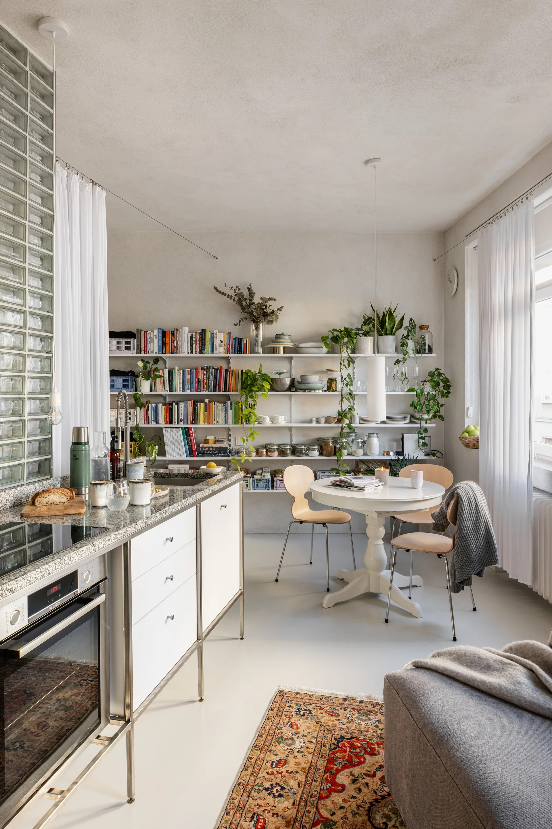 An open kitchen and dining space features a granite-topped island, a round white pedestal table with wood chairs, and wall-mounted shelving for books.