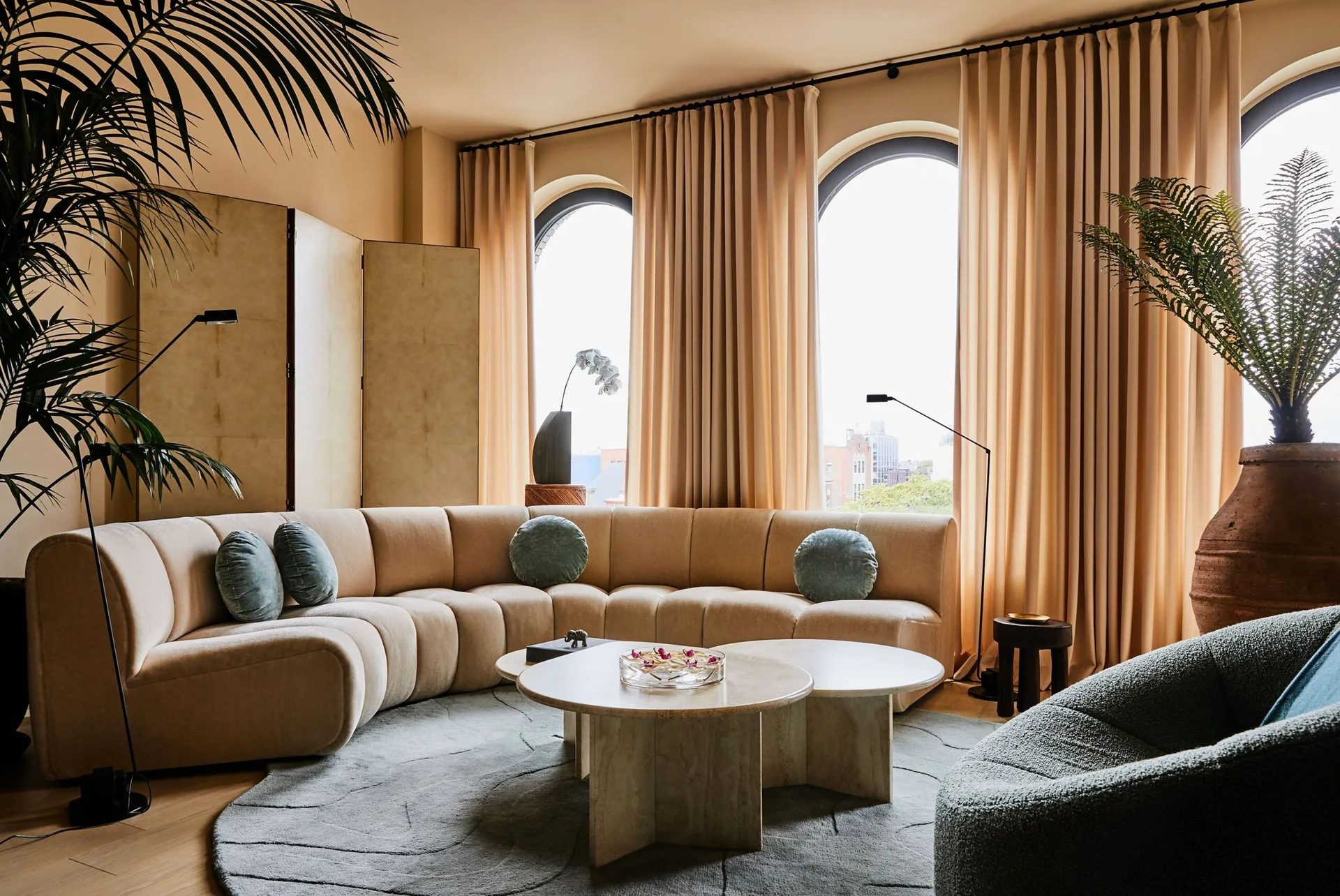 A contemporary living room featuring a curved channel-tufted beige sofa, nested stone coffee tables, and large arched windows with matching drapes.
