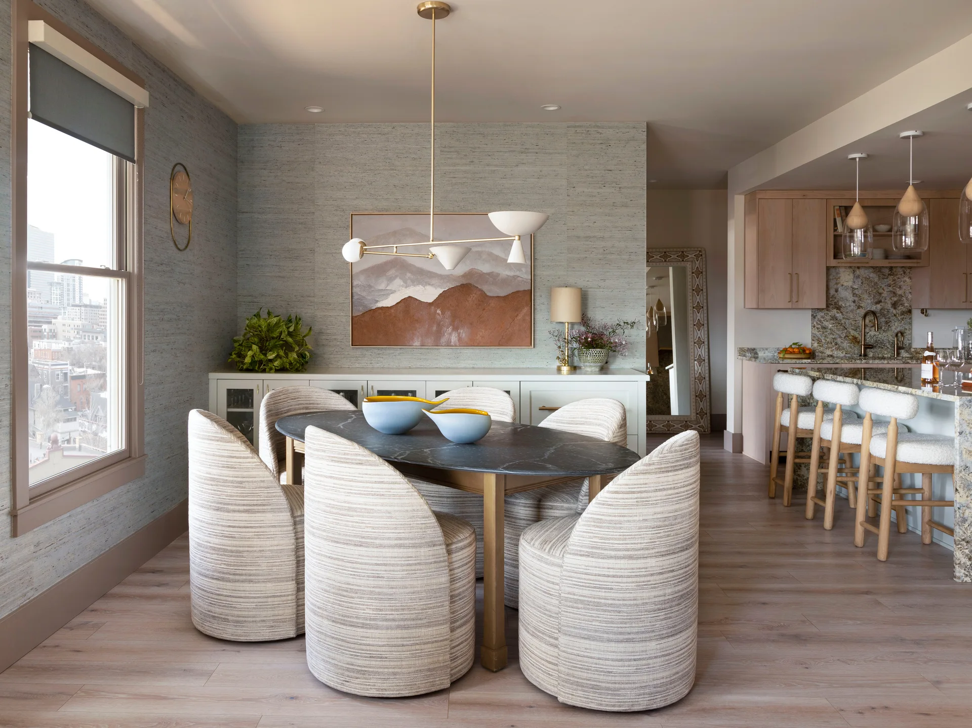 A contemporary dining room features an oval stone-topped table, curved upholstered chairs, textured gray wallpaper, and an adjacent kitchen.