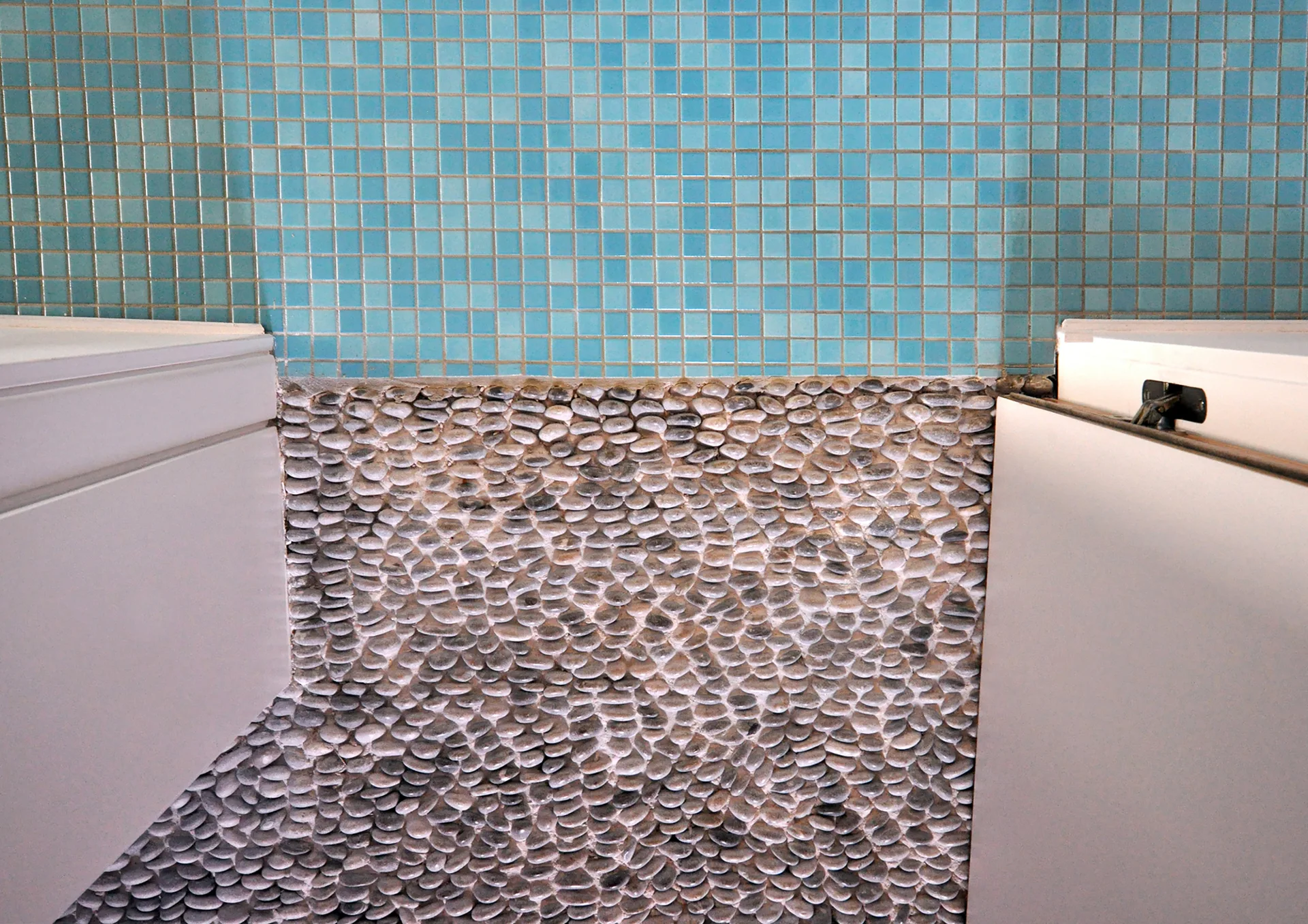 A close-up shot of a bathroom featuring a blue square mosaic tile wall above a textured grey pebble stone floor between white cabinets.