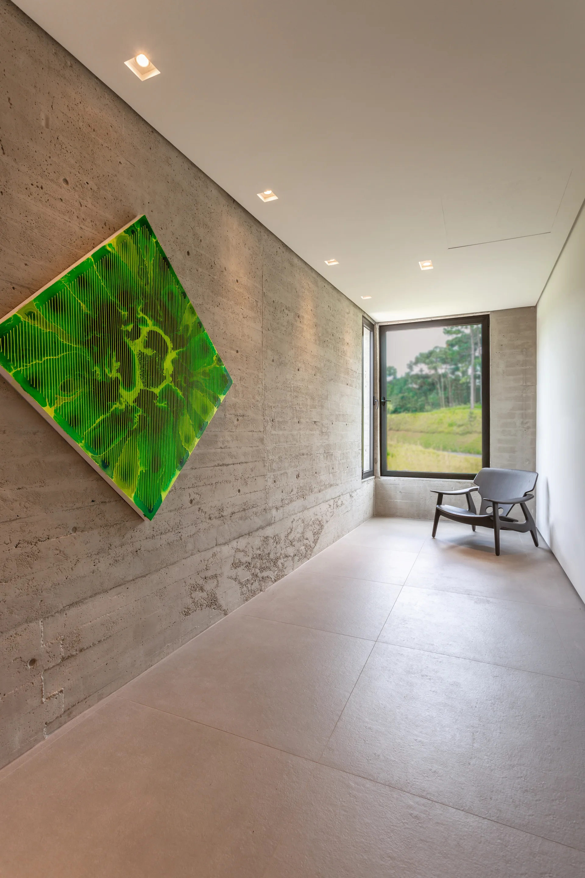 A minimalist hallway features a board-formed concrete wall with diamond-shaped green artwork and a dark lounge chair by a window.