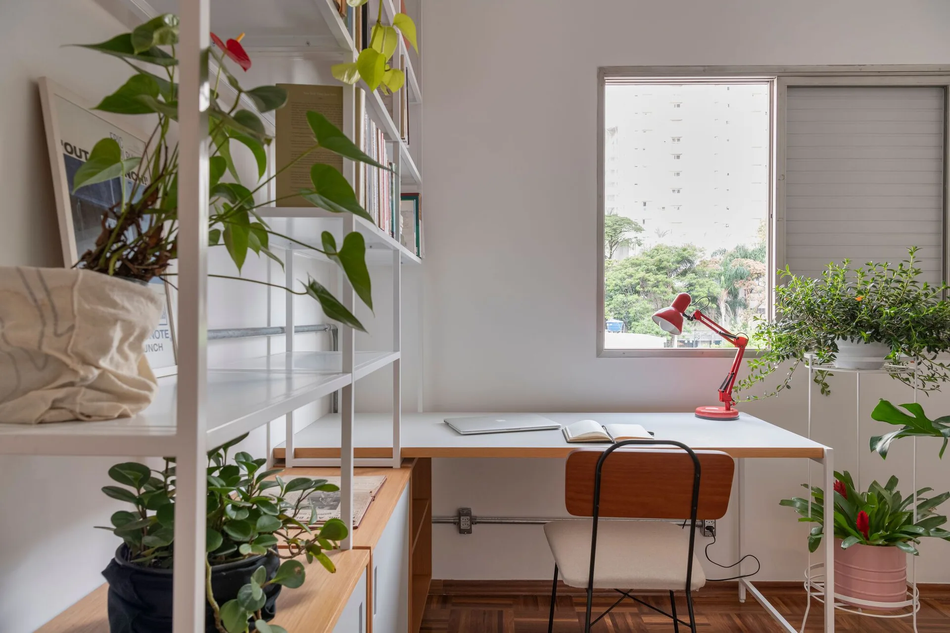 A bright home office features a white desk with a red task lamp, open shelving with plants, and a wooden chair on parquet flooring.