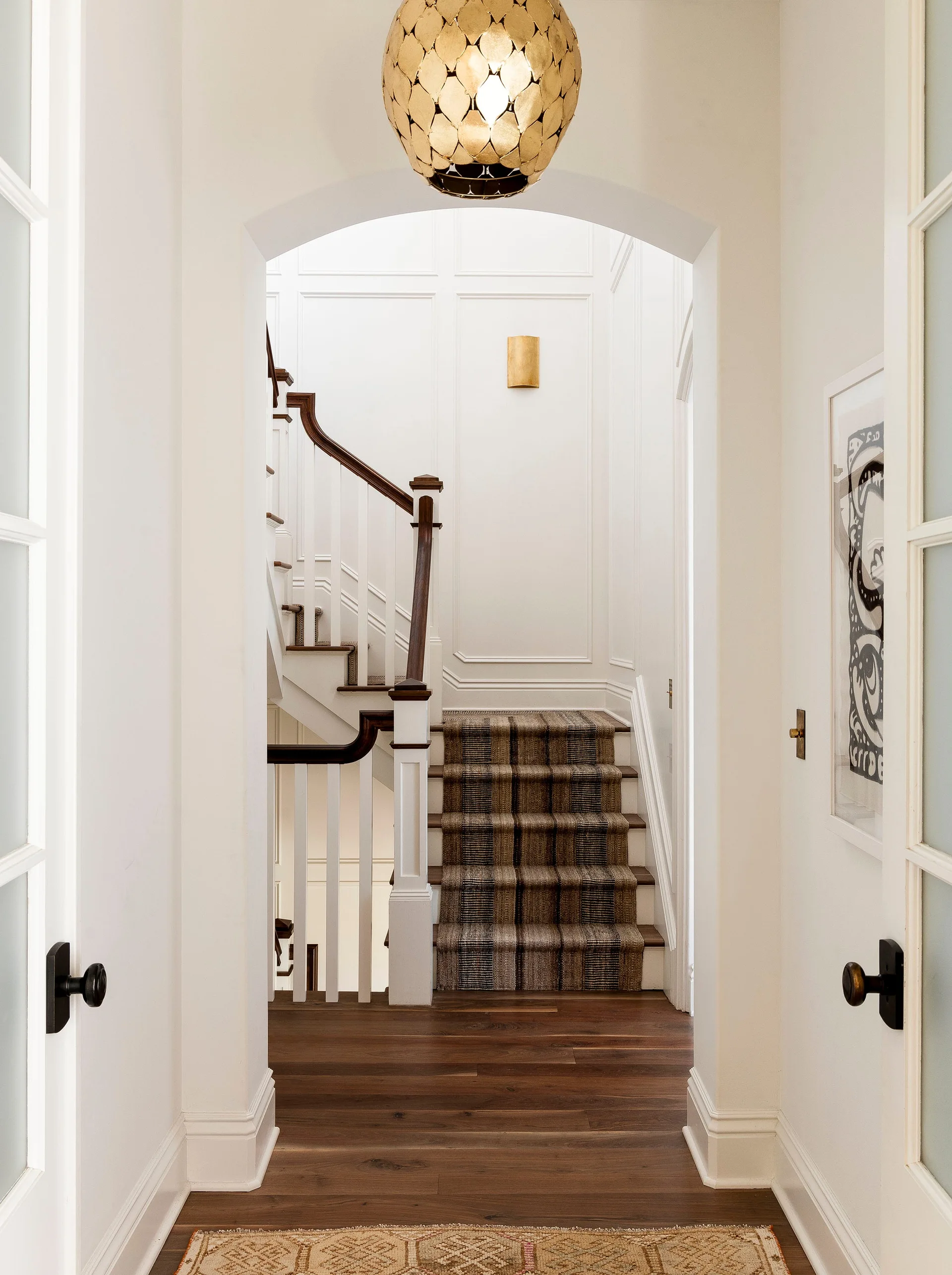 A view through a white archway showing a staircase with dark wood treads, a plaid runner, and paneled walls with a gold sconce.
