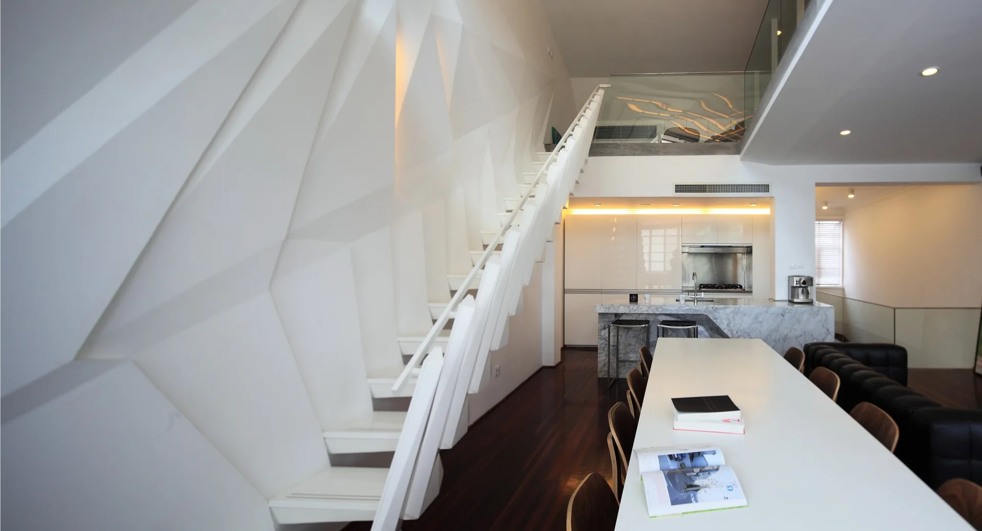 A minimalist open-plan loft featuring a sculptural geometric white wall, a floating staircase, a marble kitchen island, and dark hardwood floors.