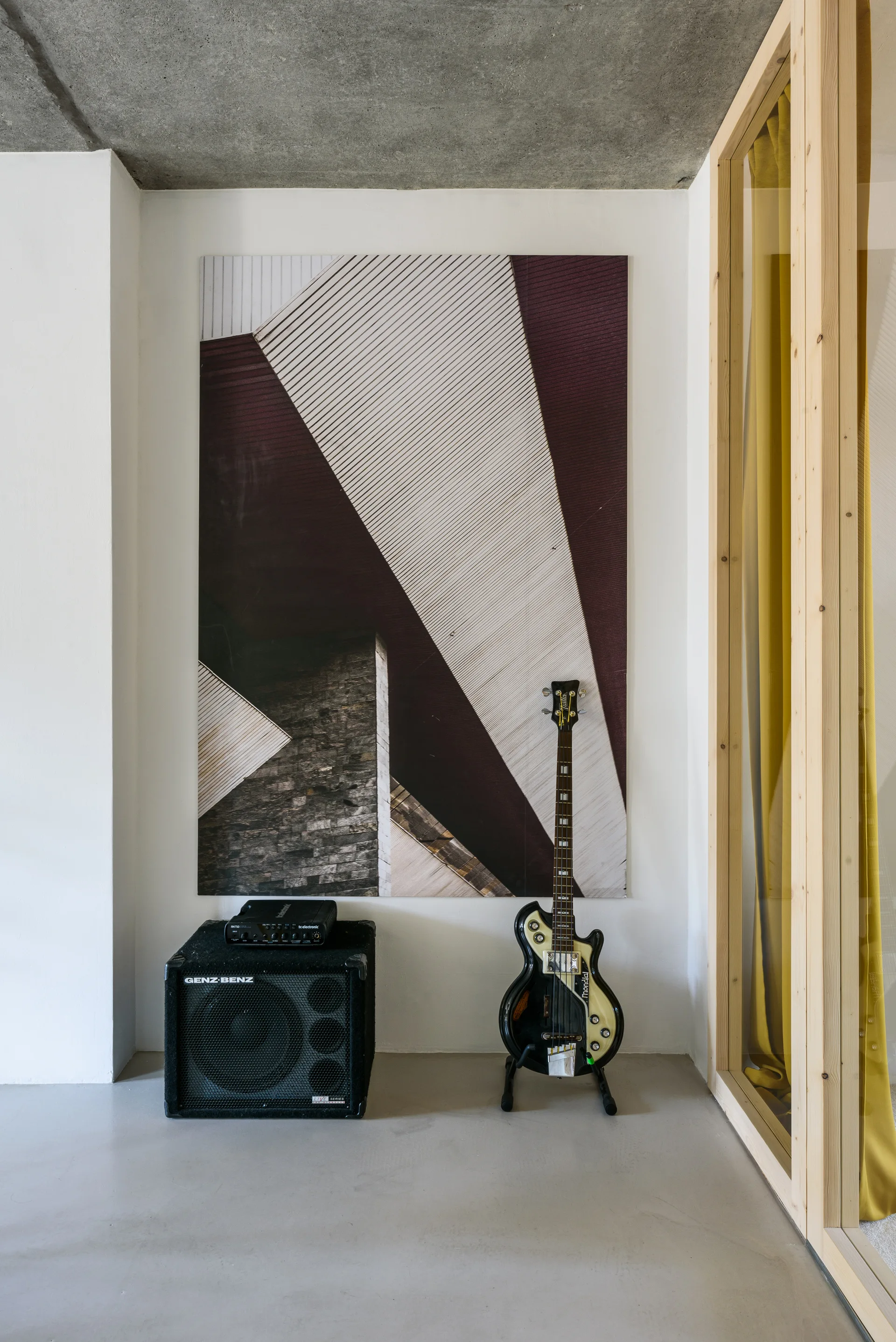 A bass guitar and amplifier sit on a grey floor beneath a large architectural print next to a light wood room divider.