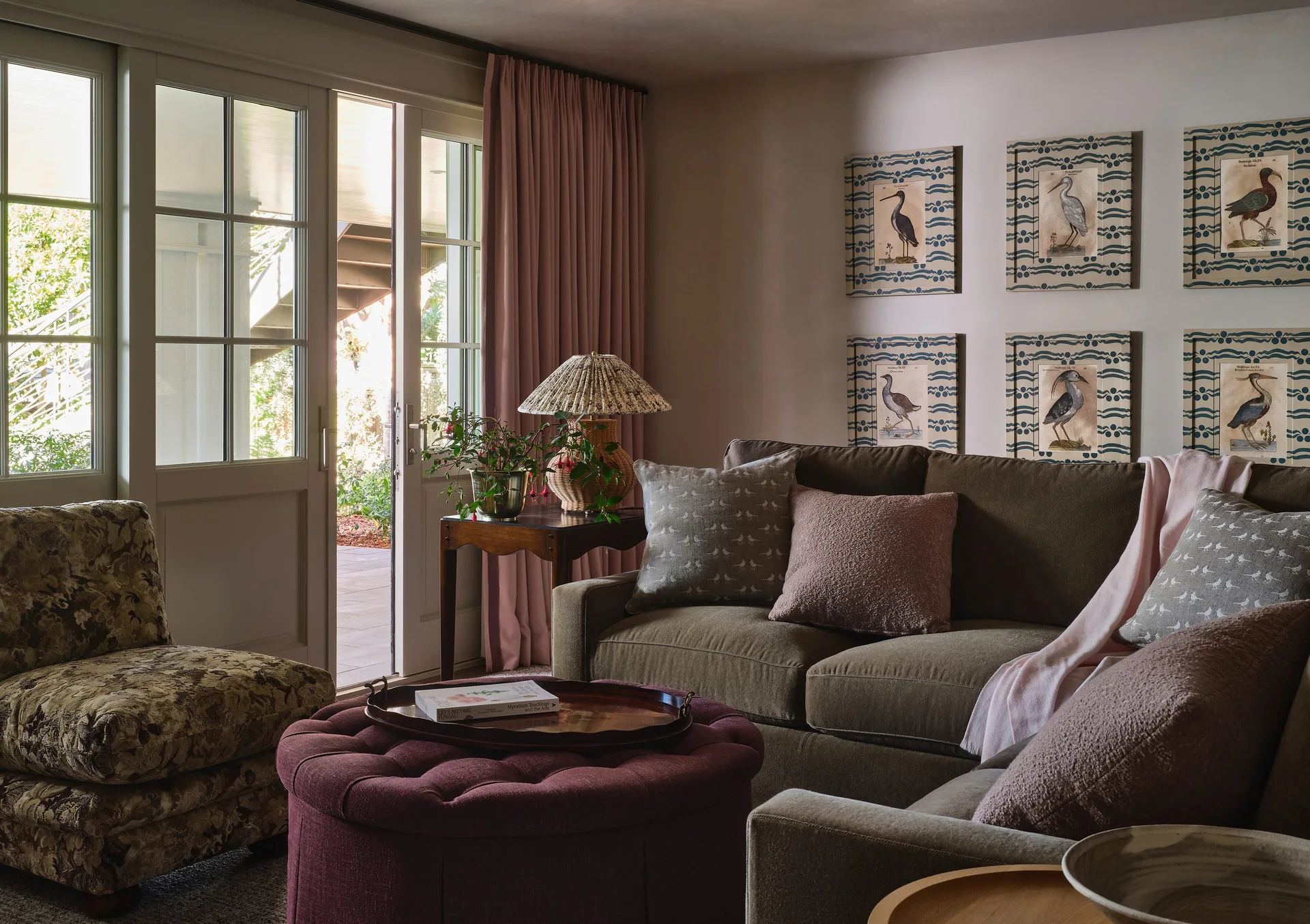 A cozy living room features an olive green sofa, a dusty pink curtain, a round plum tufted ottoman, and a grid of six framed prints on the wall.