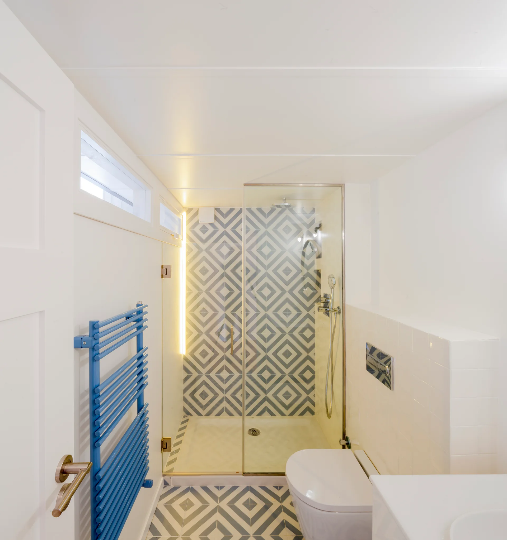 A compact modern bathroom features blue and white geometric tile on the floor and shower wall, a glass door, a wall-hung toilet, and a blue towel warmer.