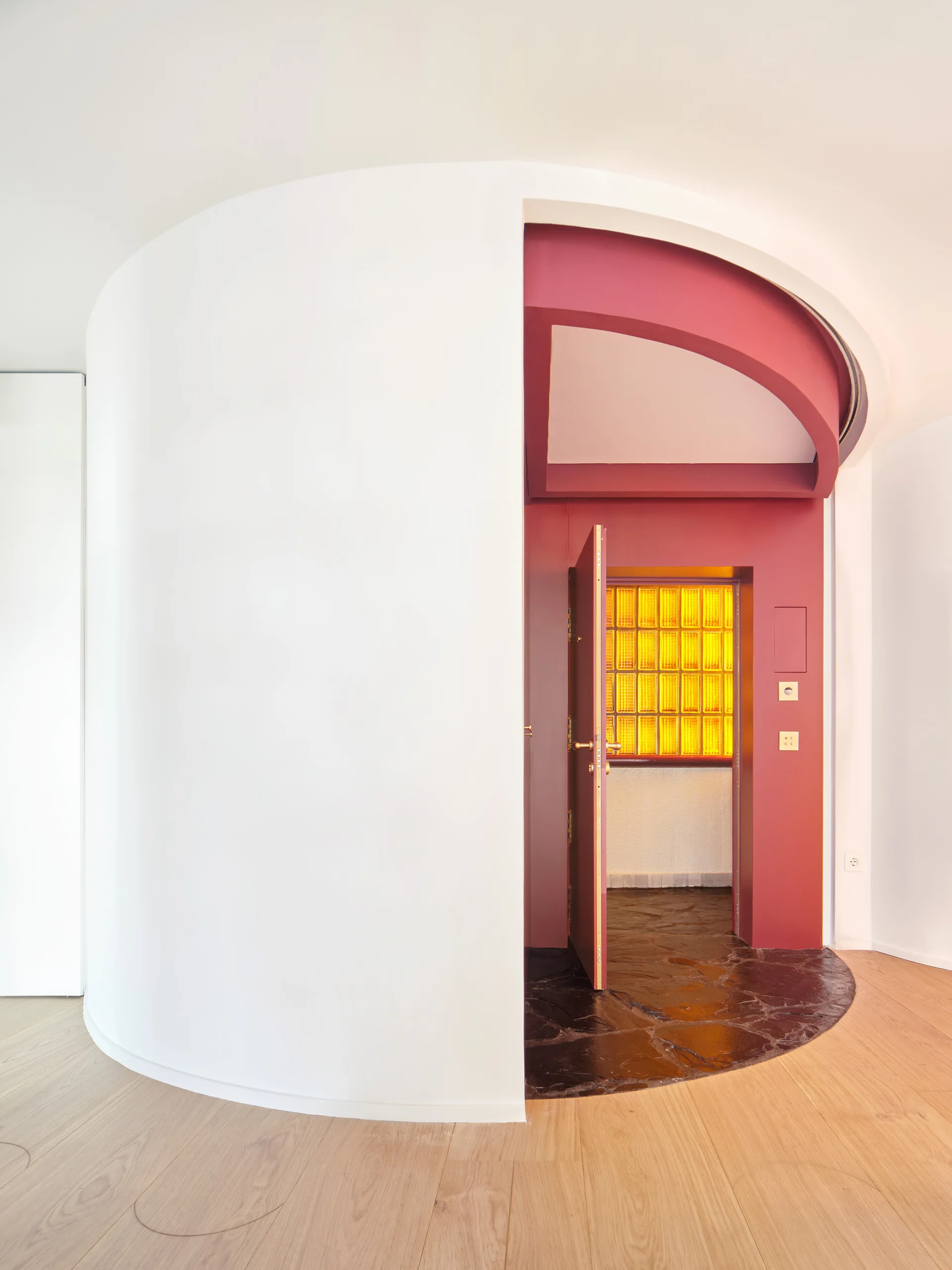 A white curved wall stands next to an open red doorway featuring a dark stone floor section and an illuminated yellow glass block wall.