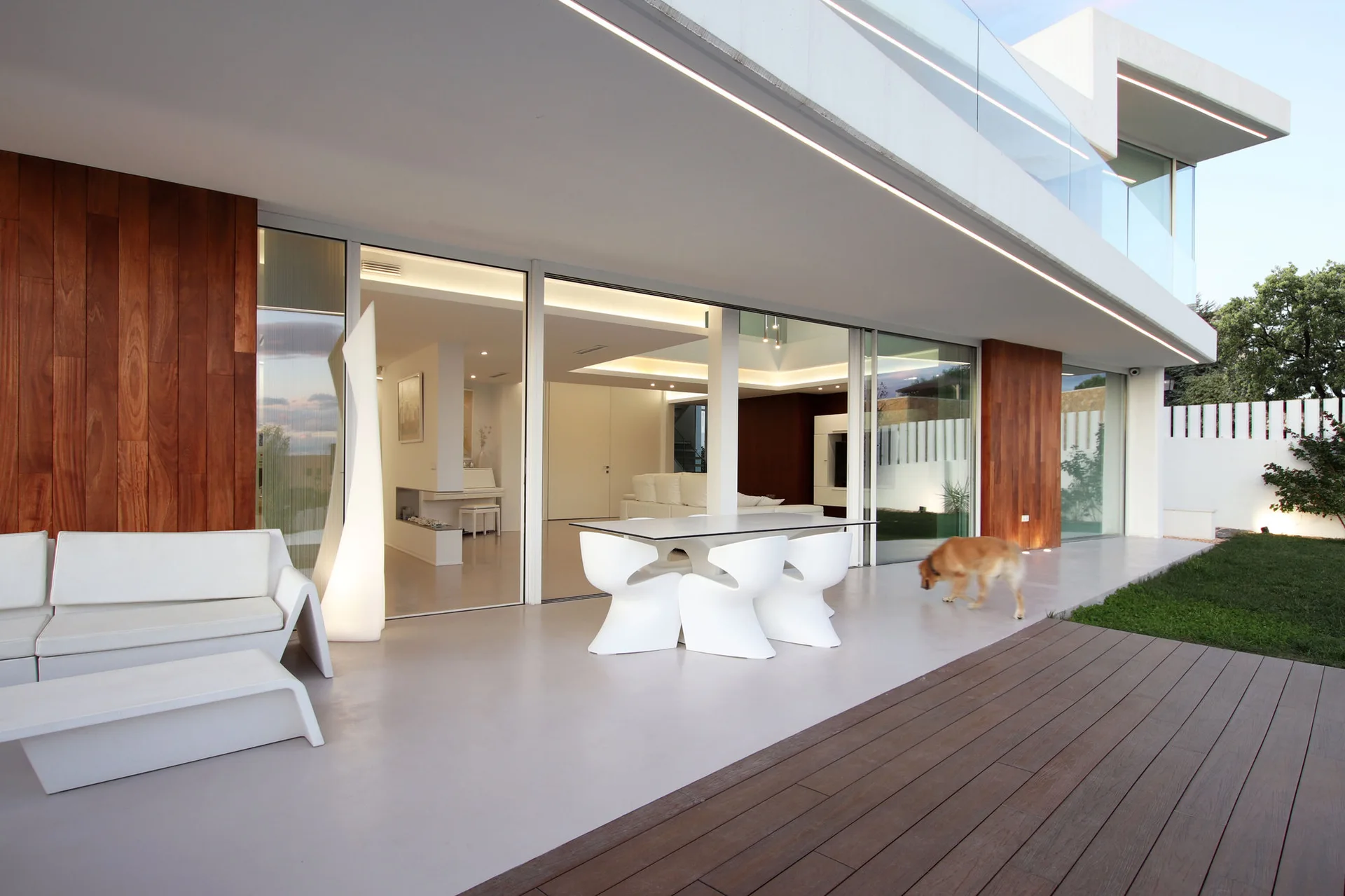 A modern minimalist patio features sculptural white dining furniture, matching outdoor sofas, and vertical wood panels alongside sliding glass doors.