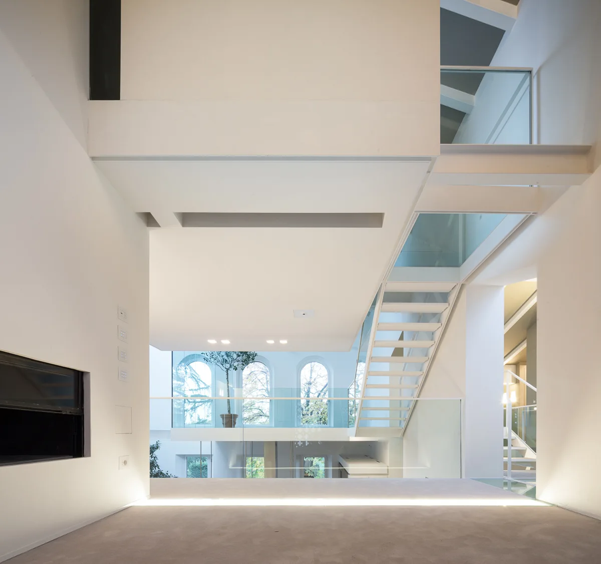 A minimalist white multi-level interior features a floating staircase with glass railings, integrated floor lighting, and distant arched windows.