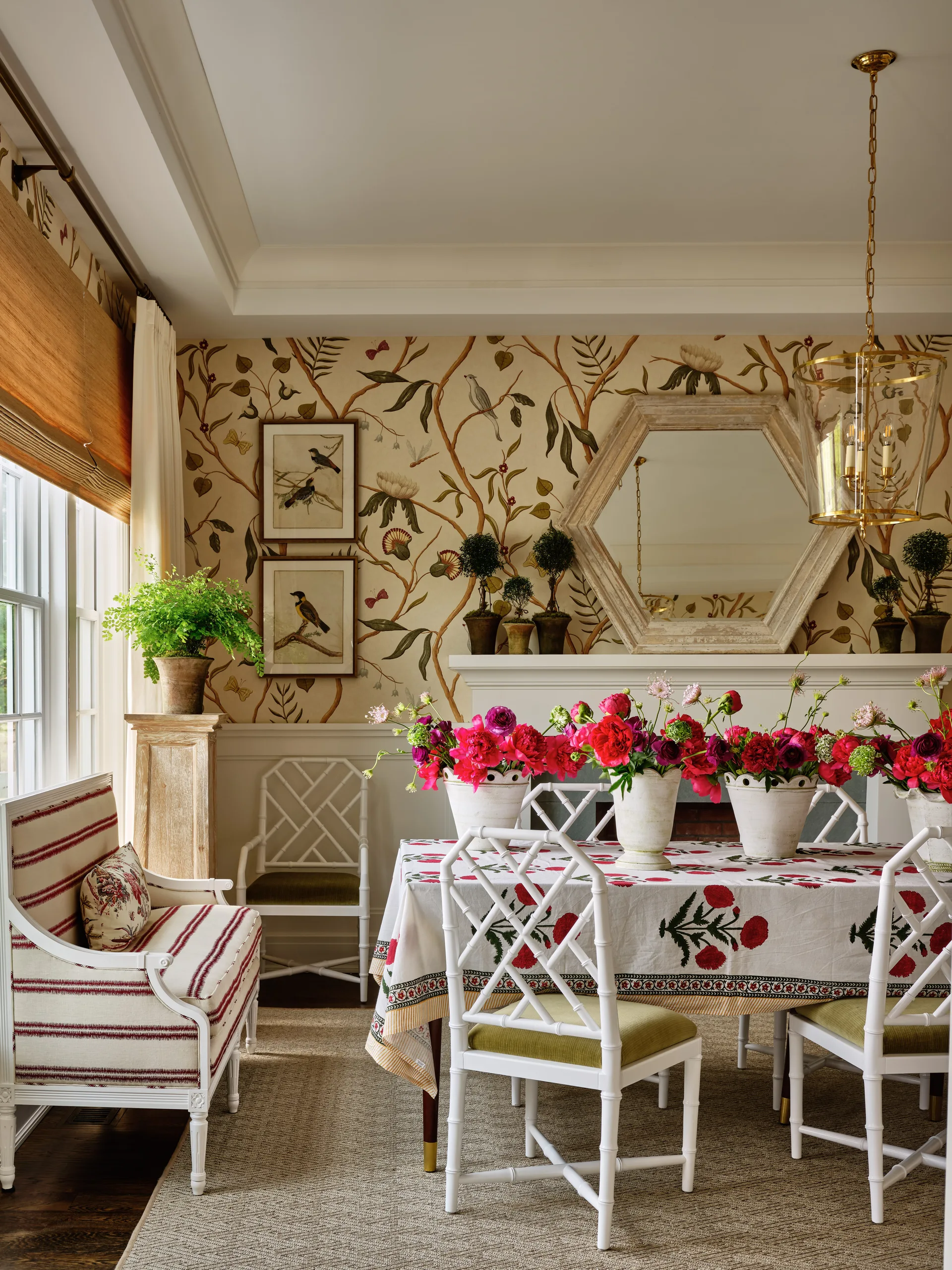 A dining room features white bamboo-style chairs around a table with a floral tablecloth, set against a backdrop of botanical wallpaper and a fireplace.