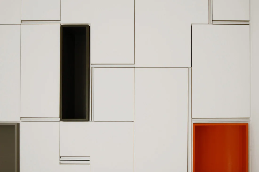 A close-up of geometric white cabinet panels with contrasting rectangular open recesses in matte black and bright orange.