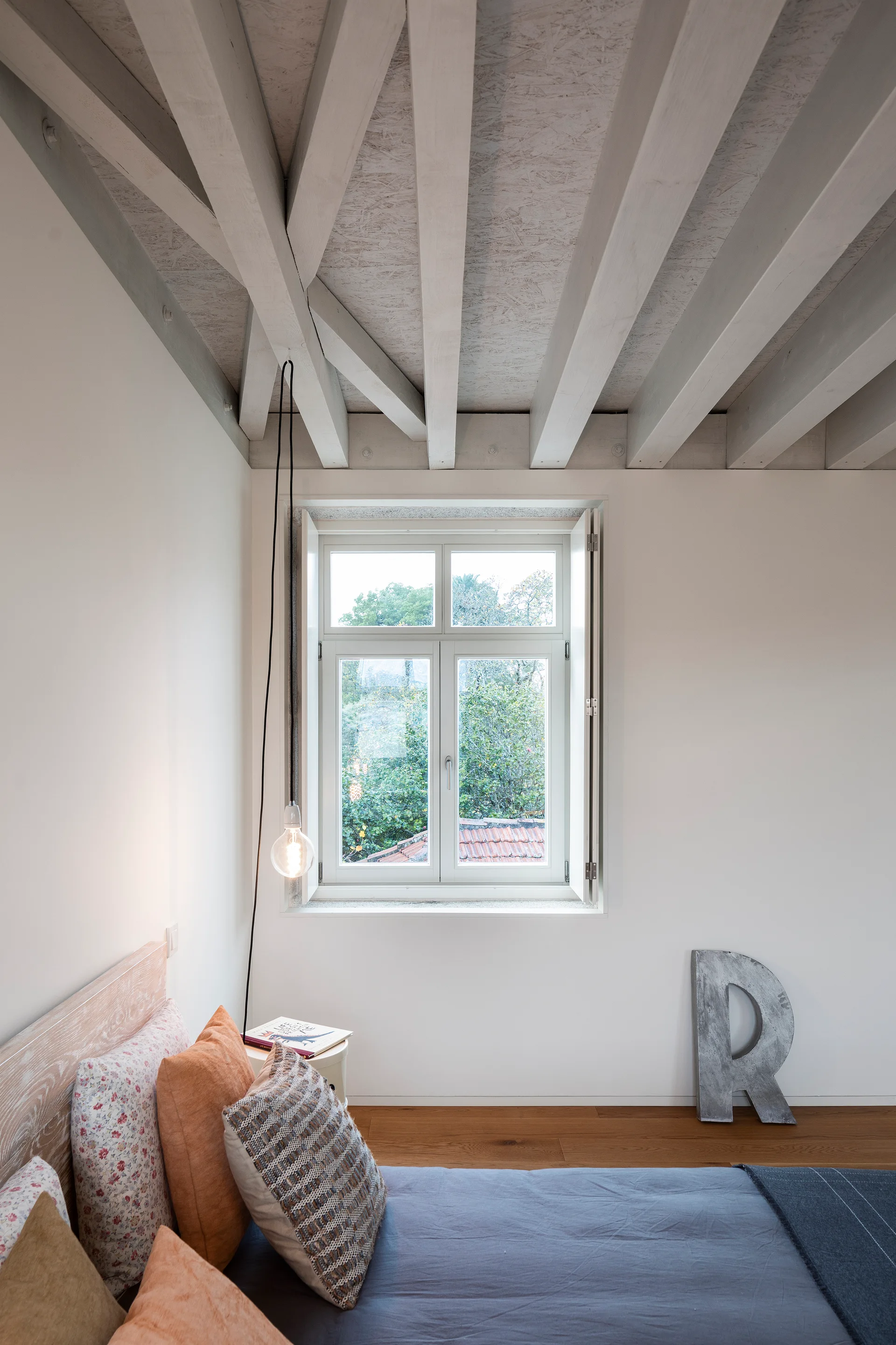 A minimalist bedroom featuring exposed white painted ceiling beams, a window looking out onto trees, and a bed with textured orange and blue bedding.