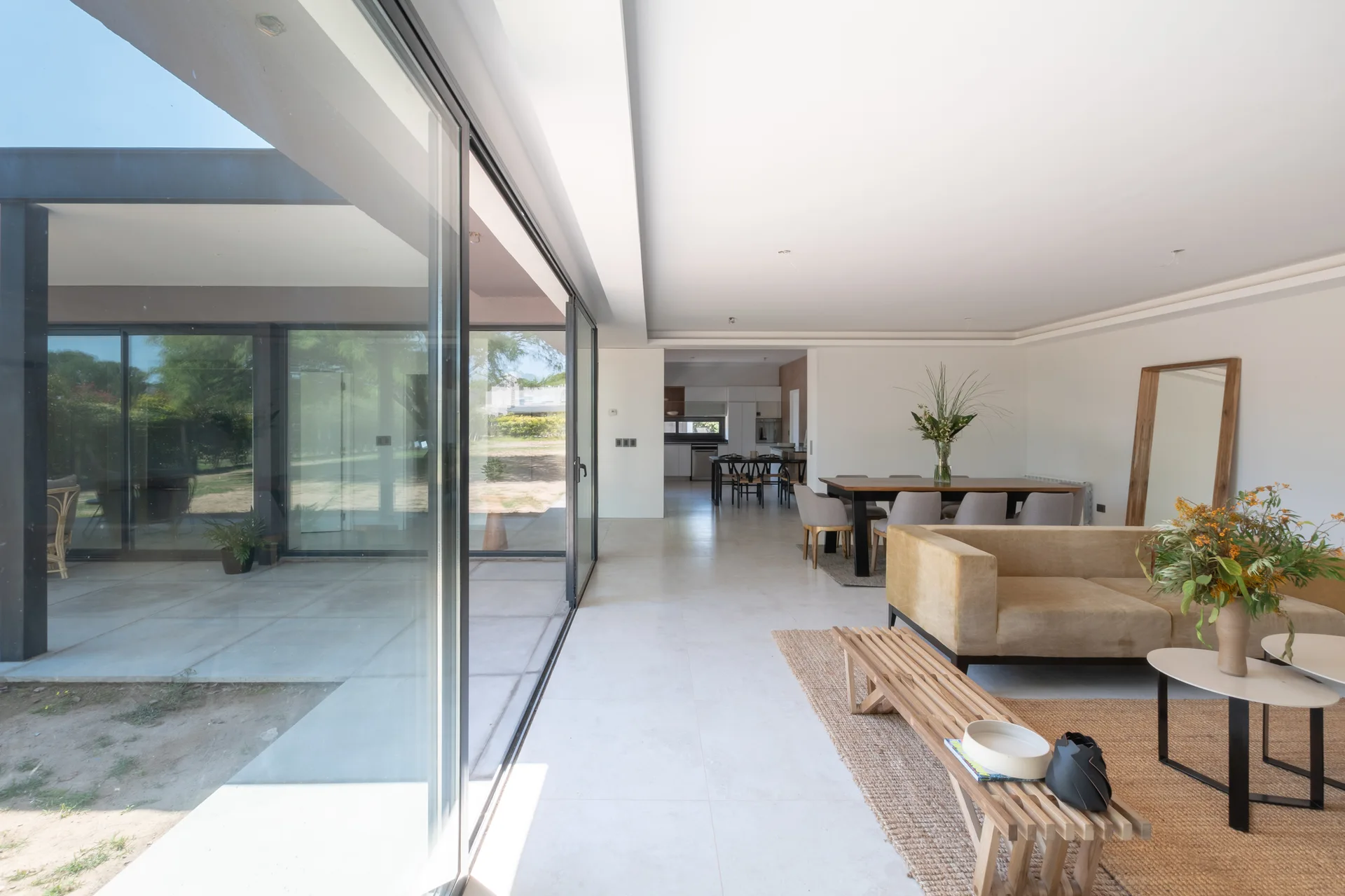 A modern open-plan living room featuring a tan sofa, slatted wooden bench, and large sliding glass doors opening to a patio.